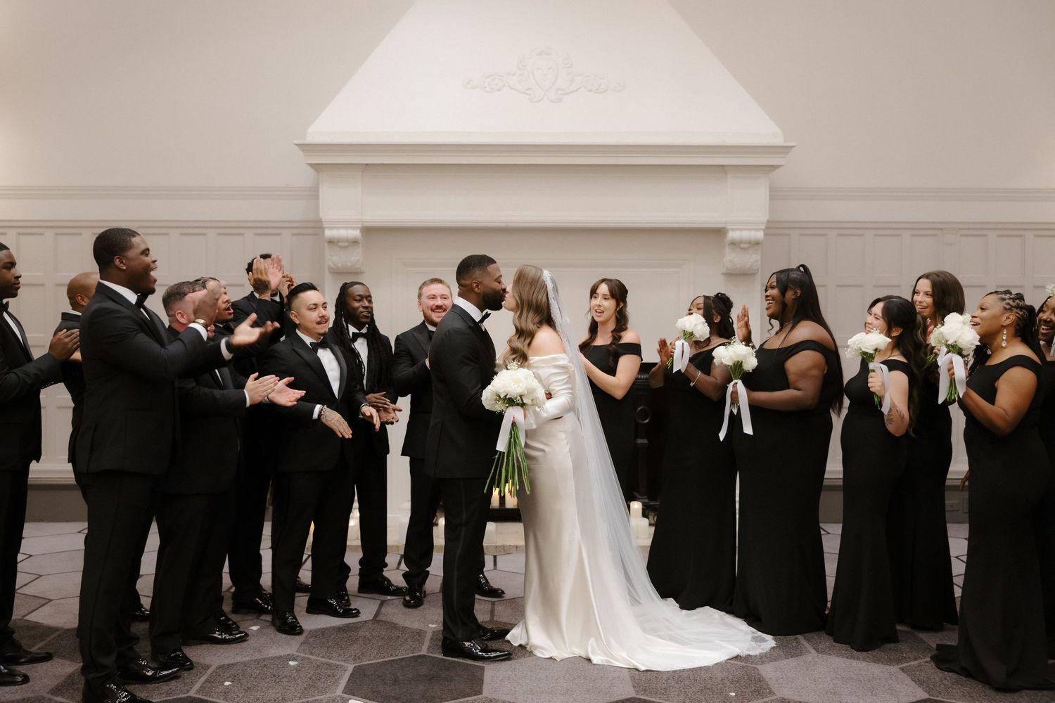 Elegant wedding party in black attire surrounds bride and groom sharing a kiss in a grand indoor venue.