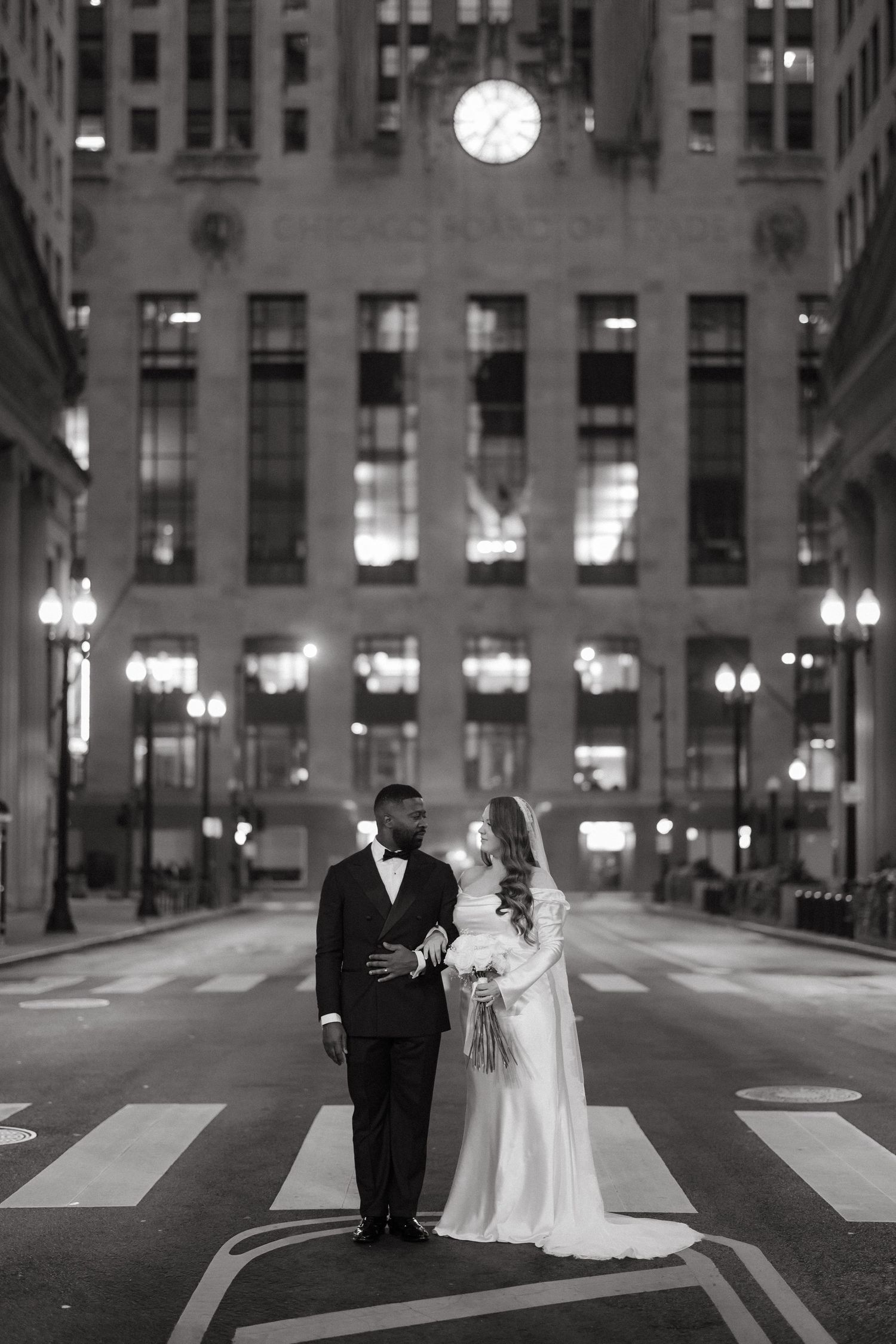 Black and white photo of a bride and groom standing on a crosswalk at night in front of a grand building with a clock.
