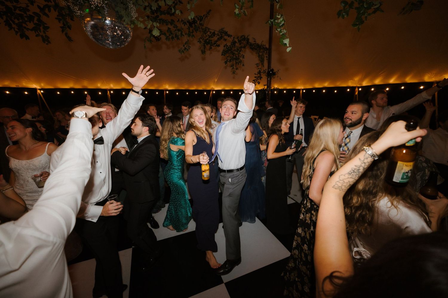 Excited wedding guests dance and celebrate with hands raised under string lights on a checkered dance floor.