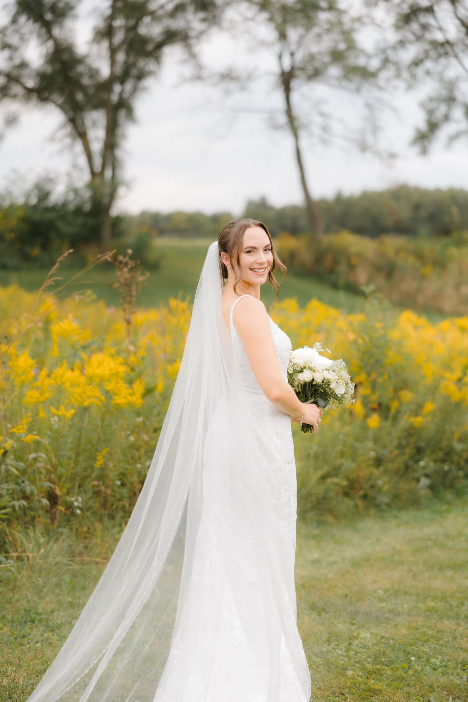 Bride in lace wedding dress with long flowing veil holding white bouquet, smiling in yellow wildflower field.