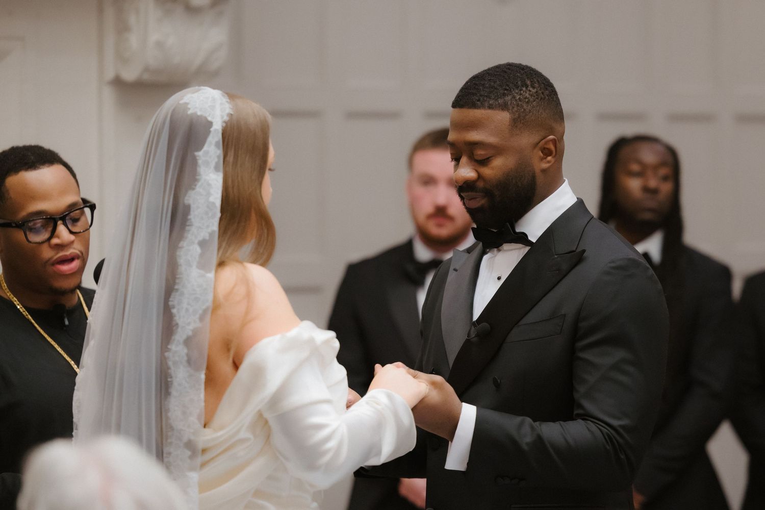 Groom in black tuxedo holds bride's hands at wedding ceremony altar, guests watching in elegant white venue.