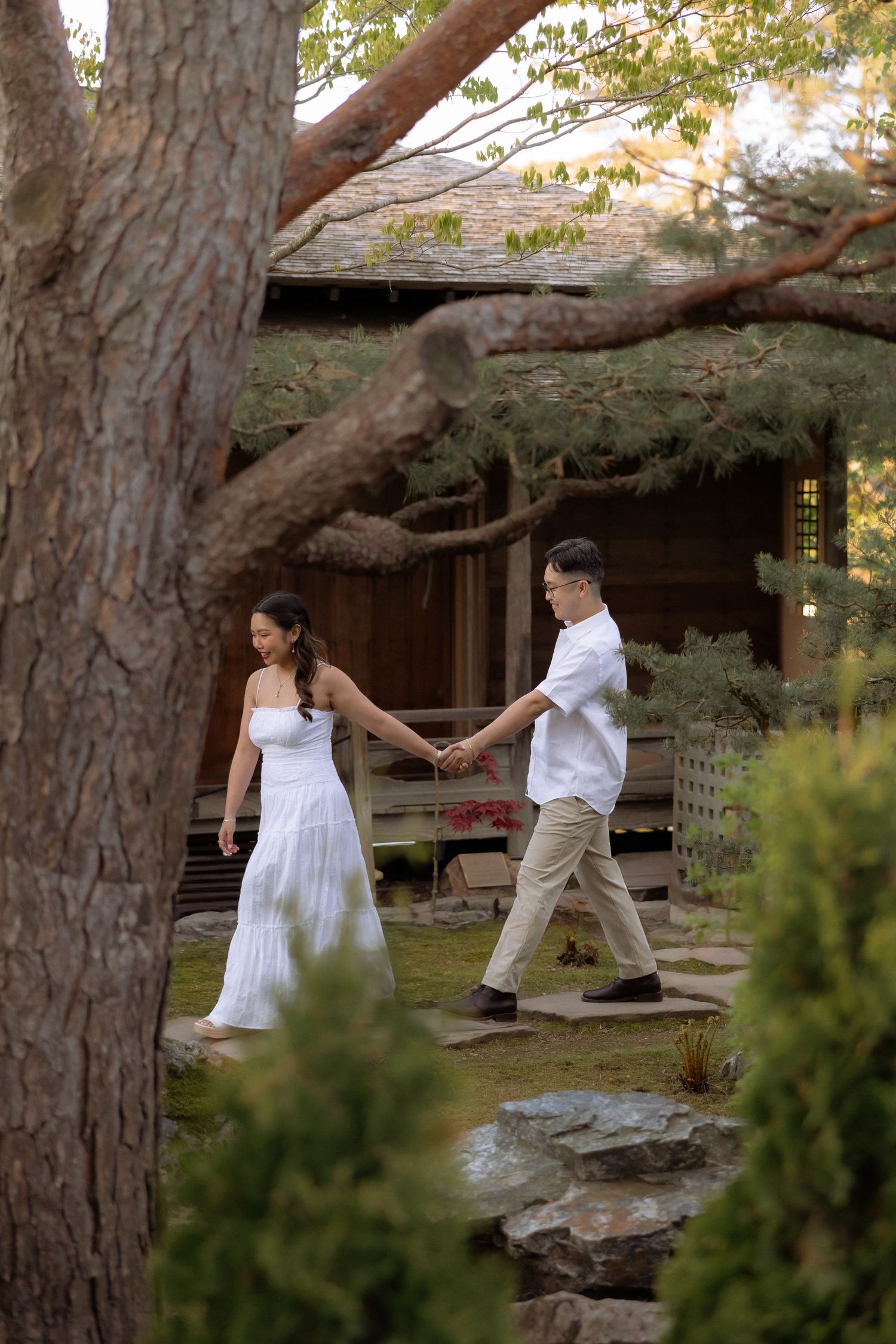 A couple in white attire holds hands while walking through a lush Japanese garden with a wooden structure in the background.