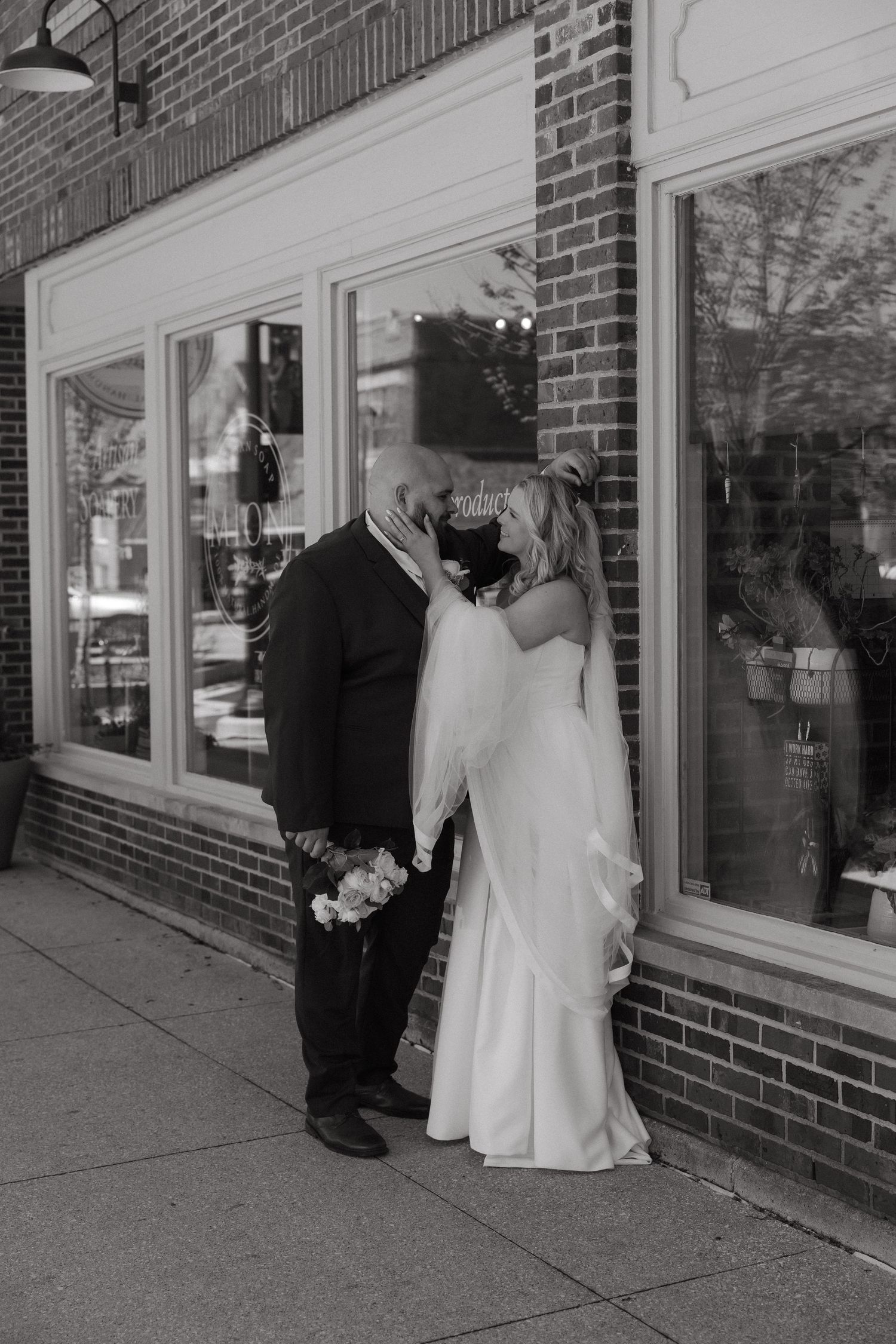 Black and white photo of a bride and groom sharing a kiss outside a brick storefront on their wedding day.