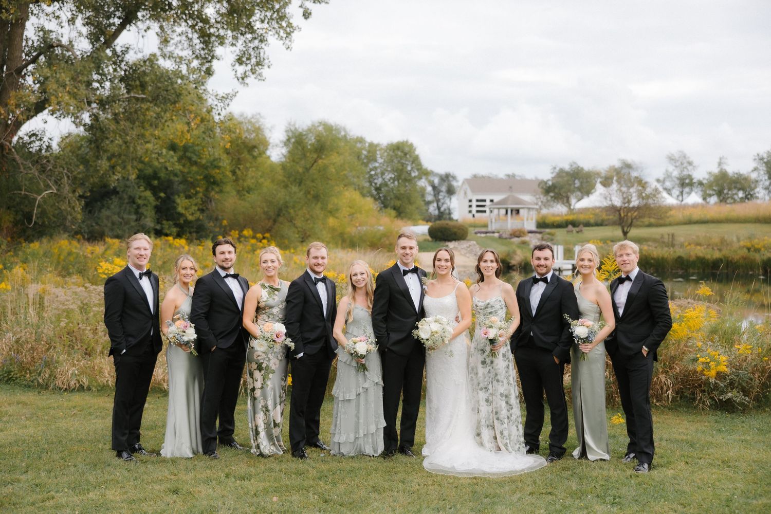 Wedding party poses formally in black tuxedos and sage floral gowns on green lawn with autumn countryside backdrop.