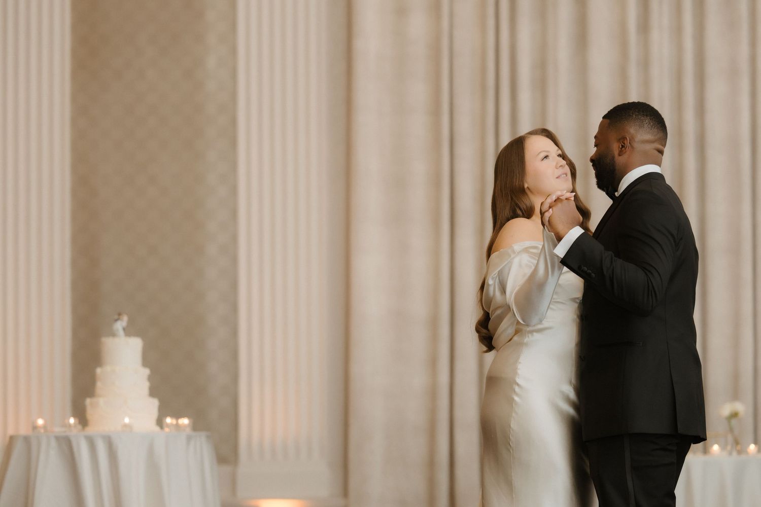 Bride and groom share first dance in elegant ballroom with white wedding cake and candles in background.