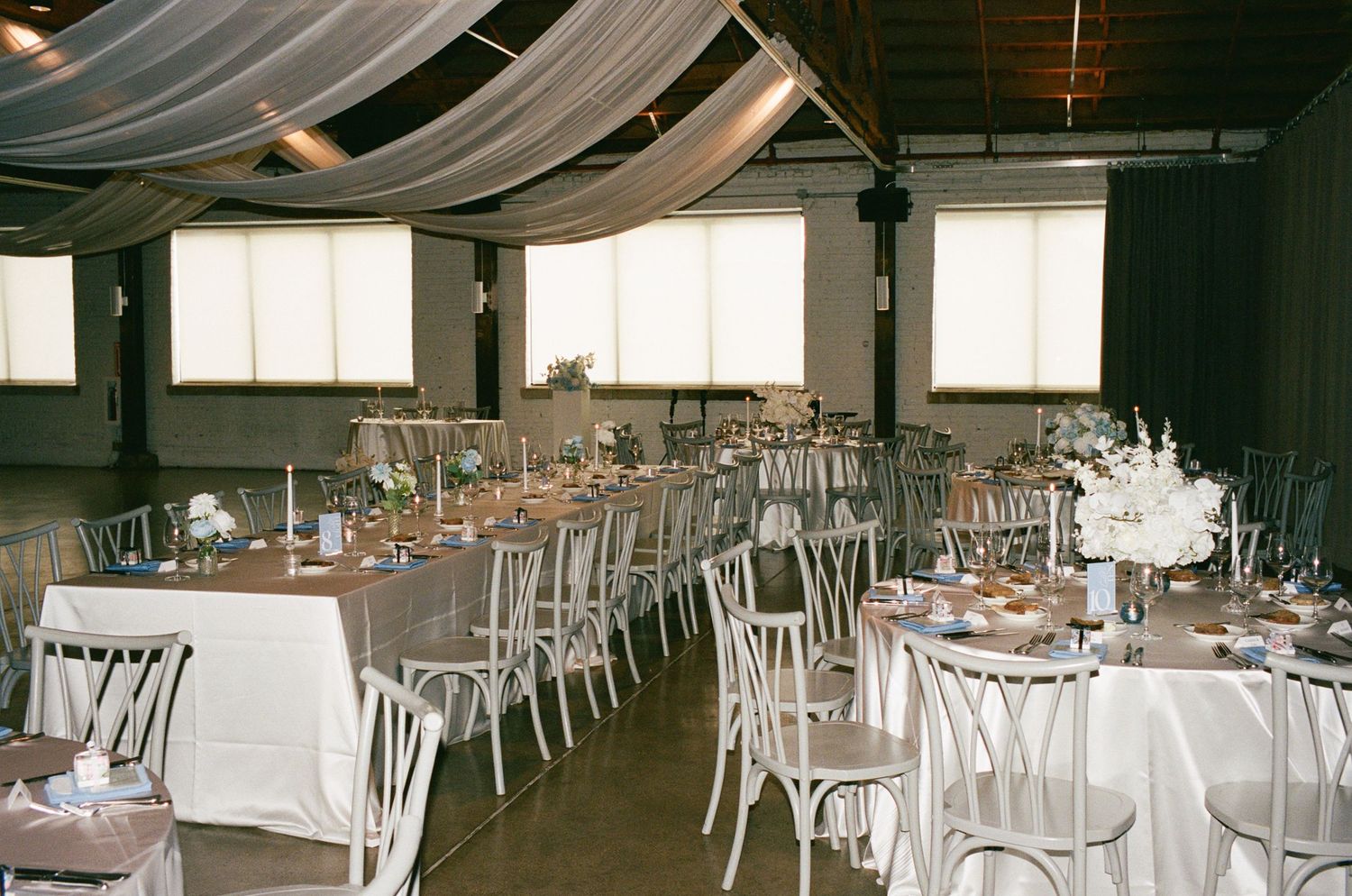Elegant wedding reception hall with long tables, white chairs, draped ceiling, and floral centerpieces in a rustic venue.