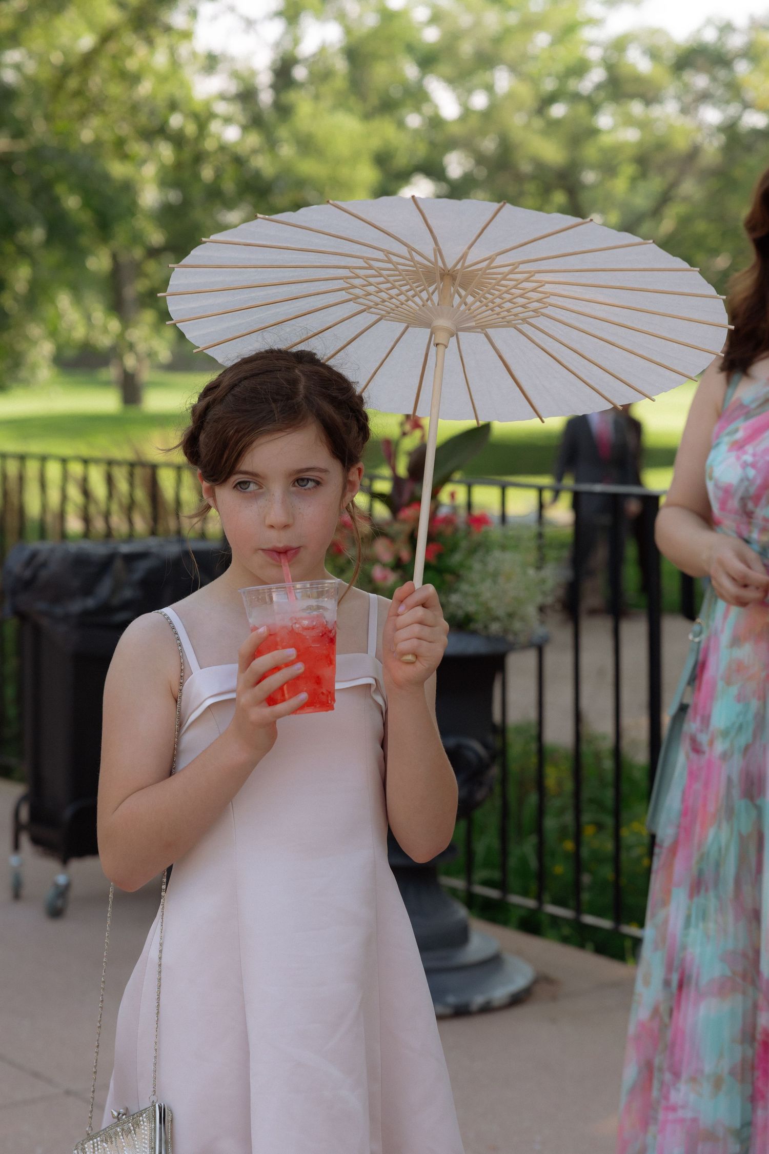 Young flower girl in pink dress sips a red drink while holding a white parasol umbrella at an outdoor wedding.