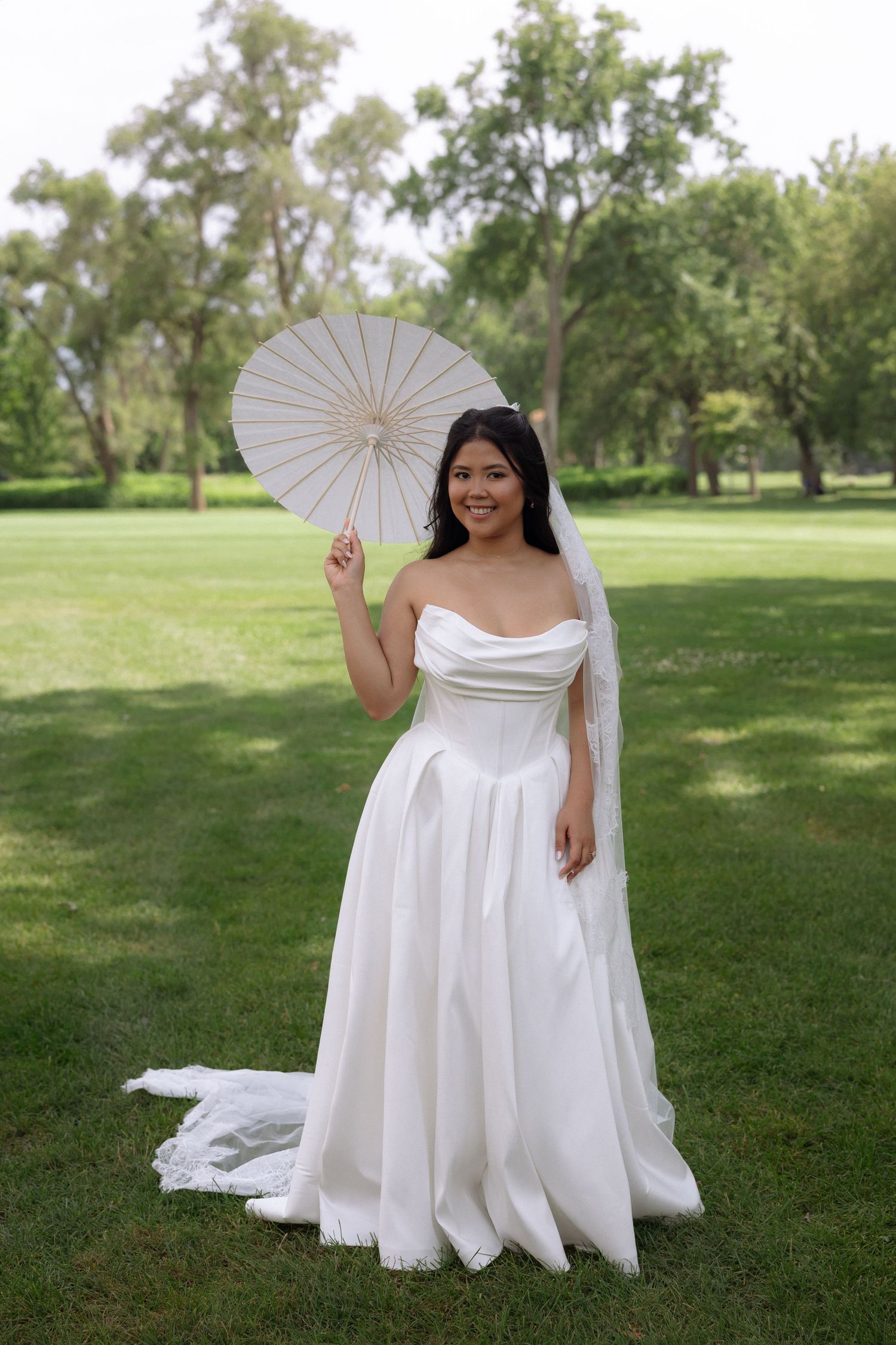 Smiling bride in a strapless white wedding gown holding a white paper parasol umbrella in a green park.