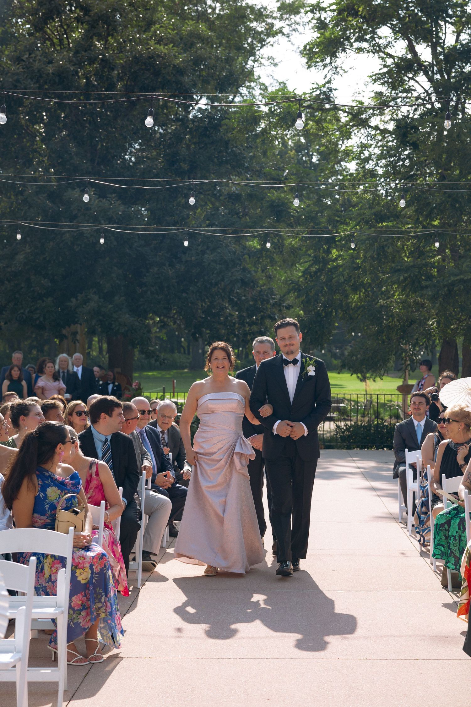 Couple walks down outdoor wedding aisle lined with seated guests and string lights amid lush trees.