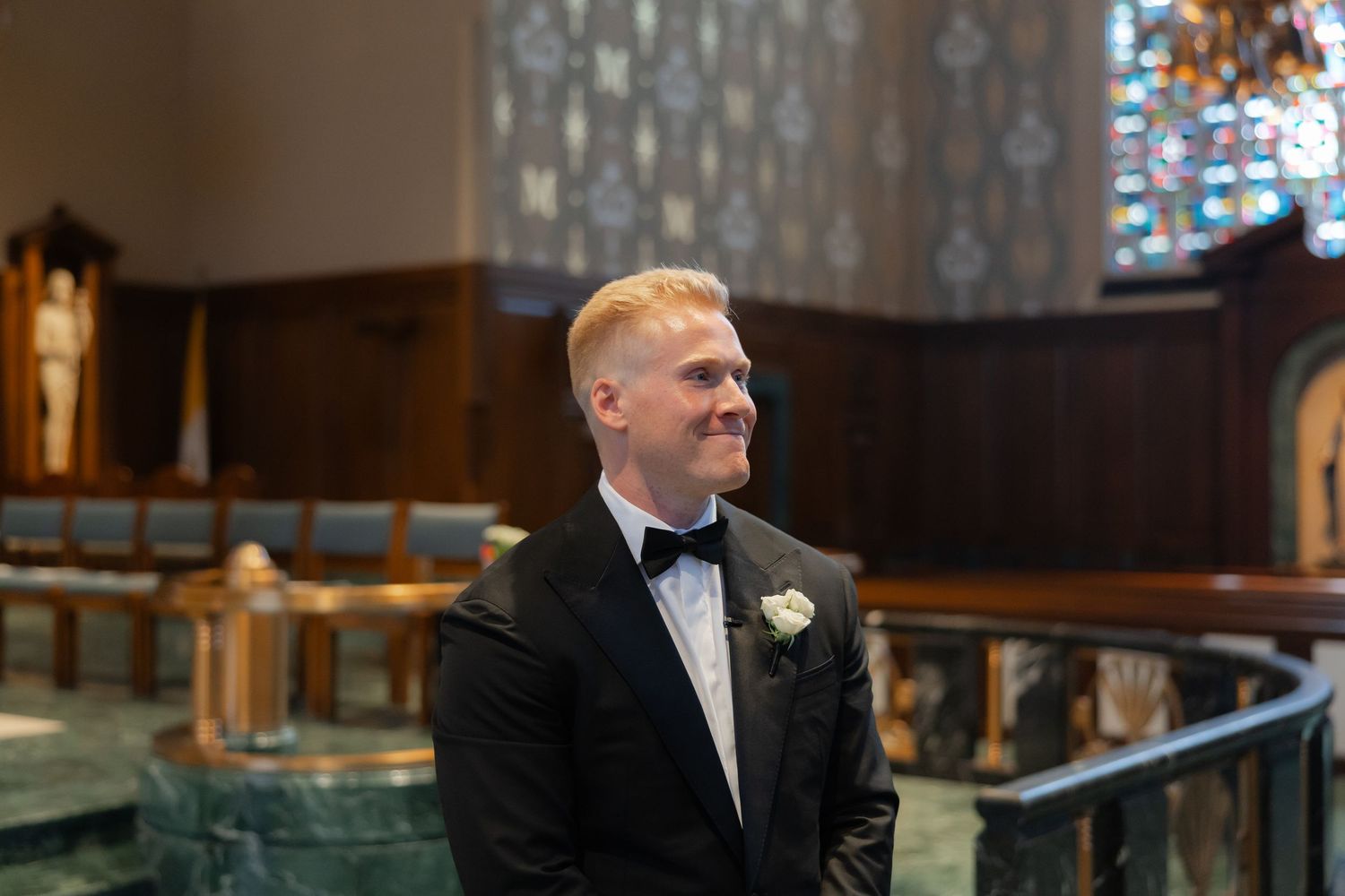 Smiling groom in black tuxedo standing at church altar awaiting bride.
