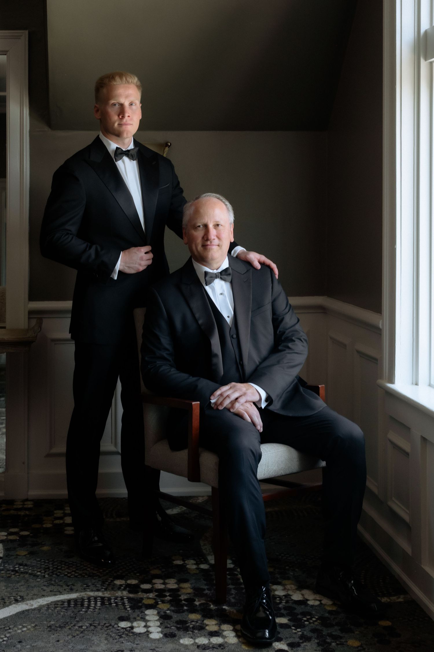 Two men in black tuxedos pose elegantly, one seated and one standing, in a classic interior setting.
