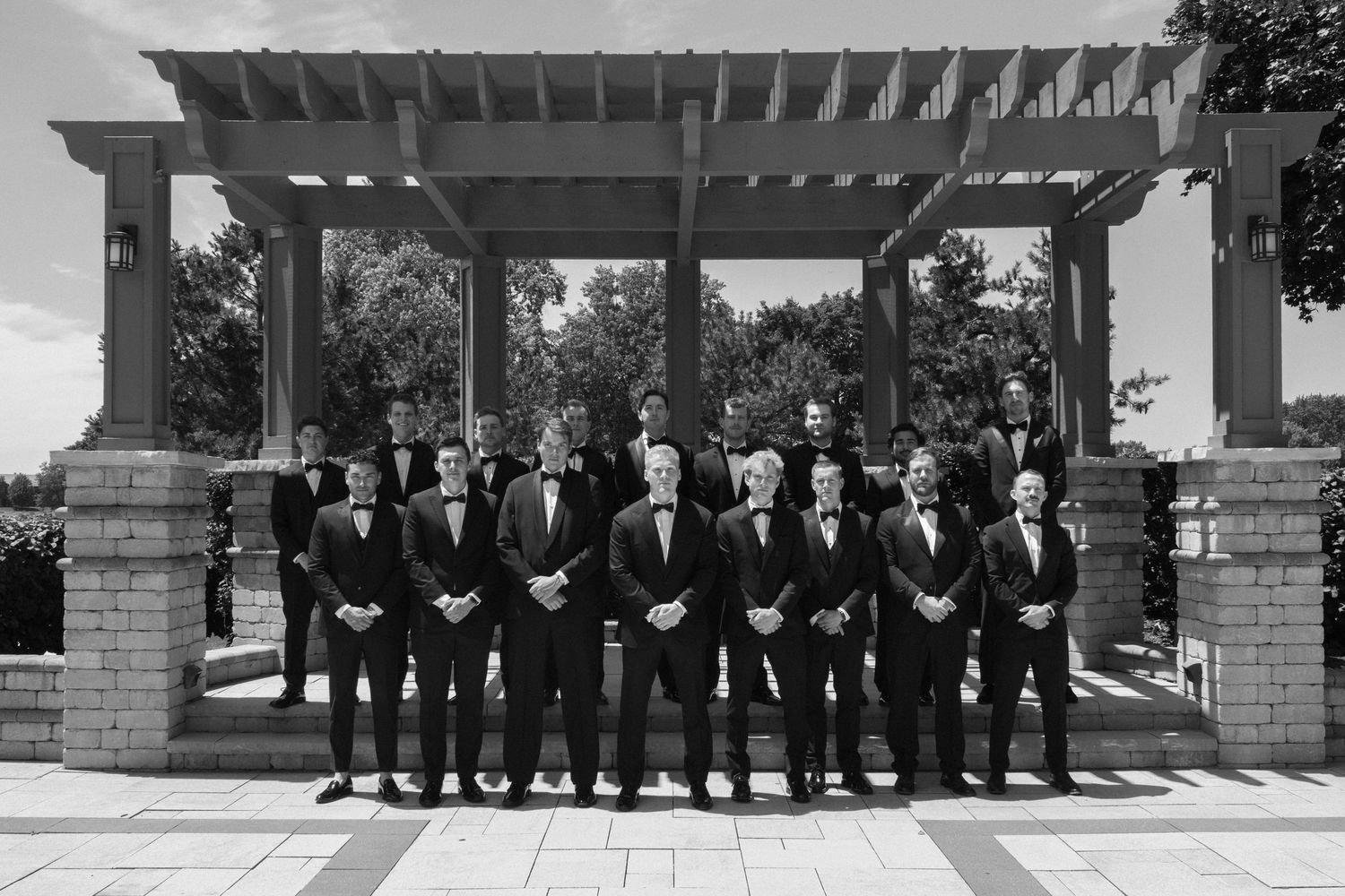 Black and white photo of groomsmen in tuxedos posing under a pergola at an outdoor wedding venue.
