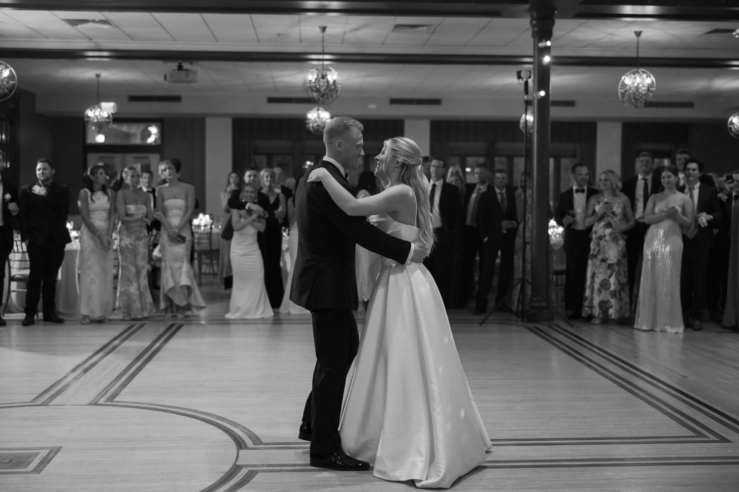 Black and white photo of bride and groom sharing first dance in grand ballroom with guests watching.
