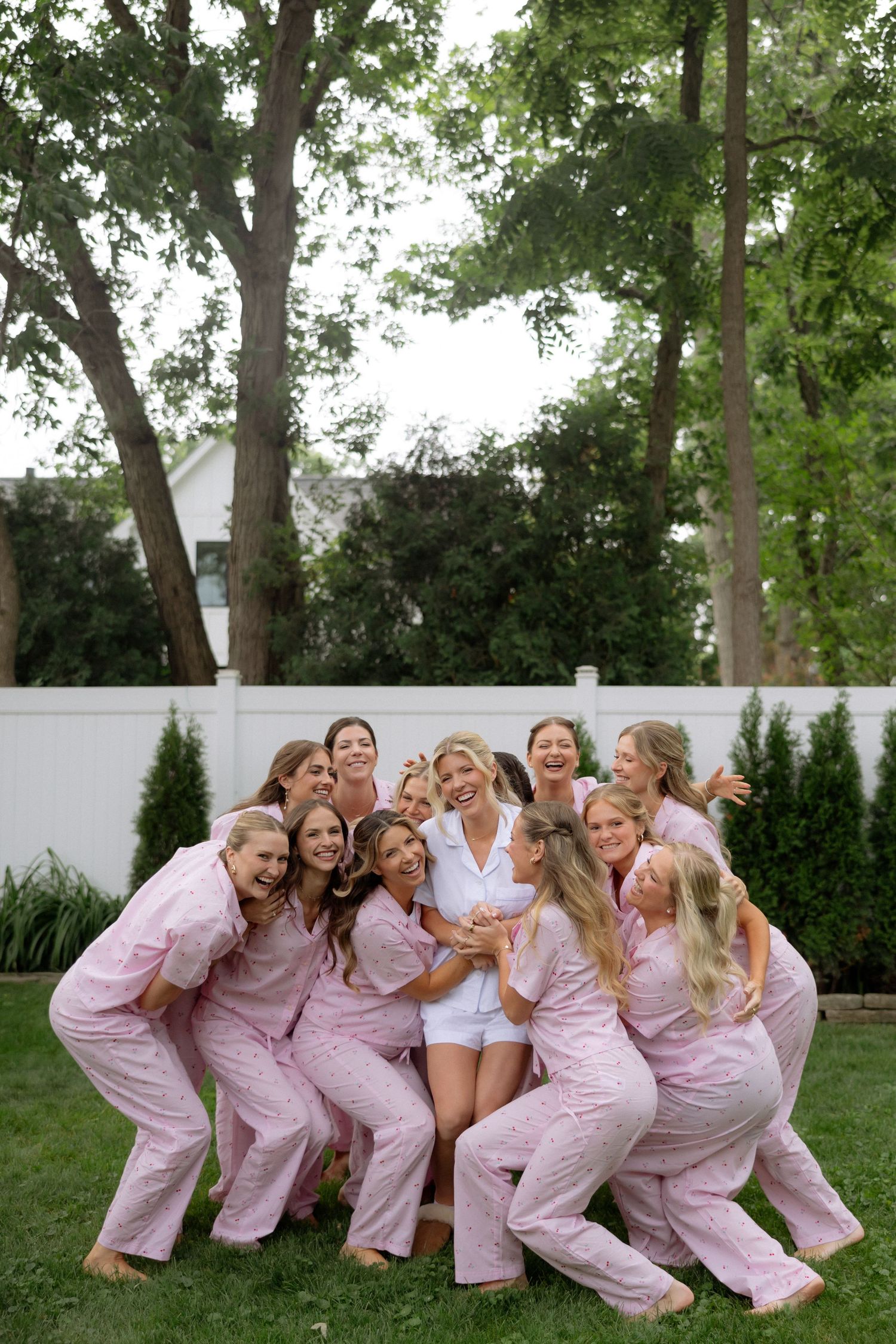 Bride in white surrounded by bridesmaids in matching pink pajamas, laughing together outdoors in a backyard.