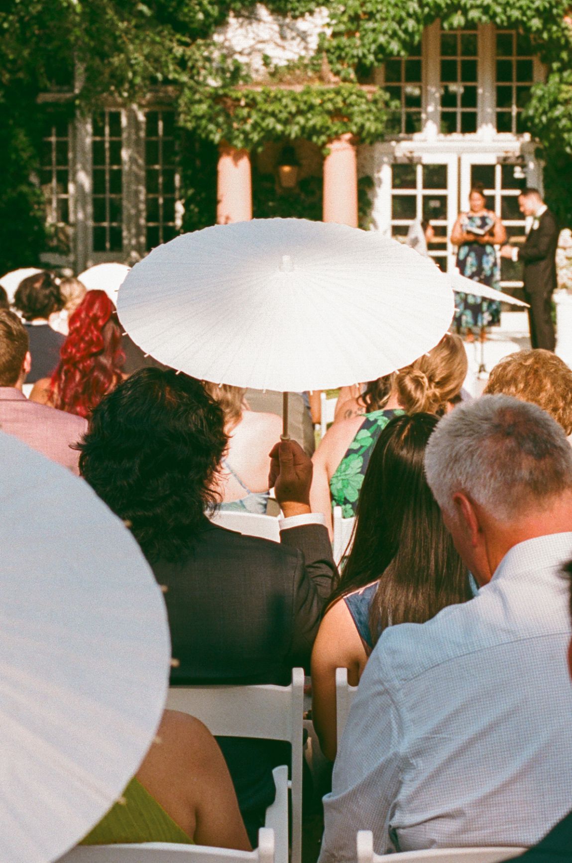 Outdoor wedding ceremony guests seated in white chairs, shielding from sun with white parasols, lush greenery backdrop.