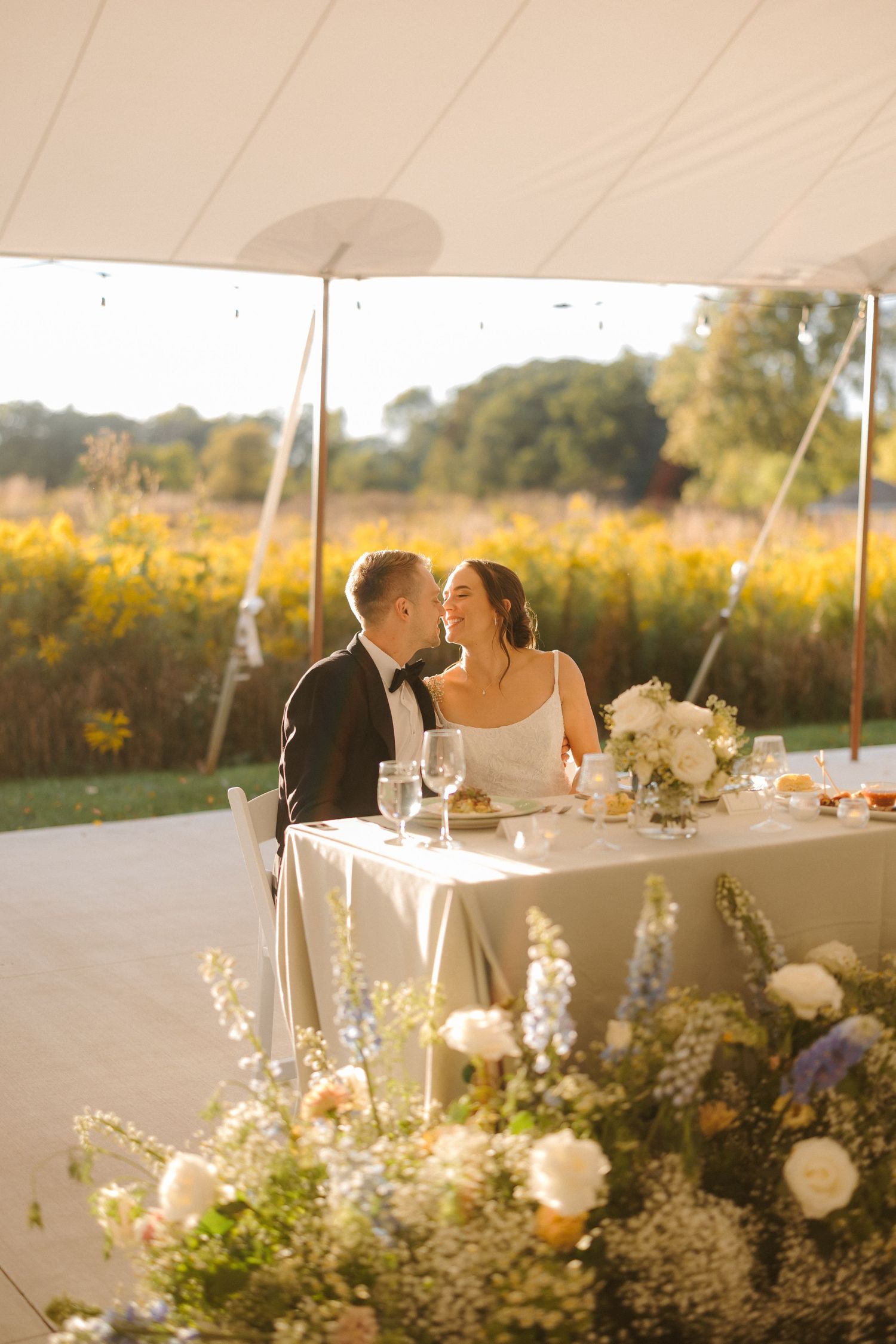 Bride and groom share a romantic moment at sweetheart table under tent with golden field backdrop.