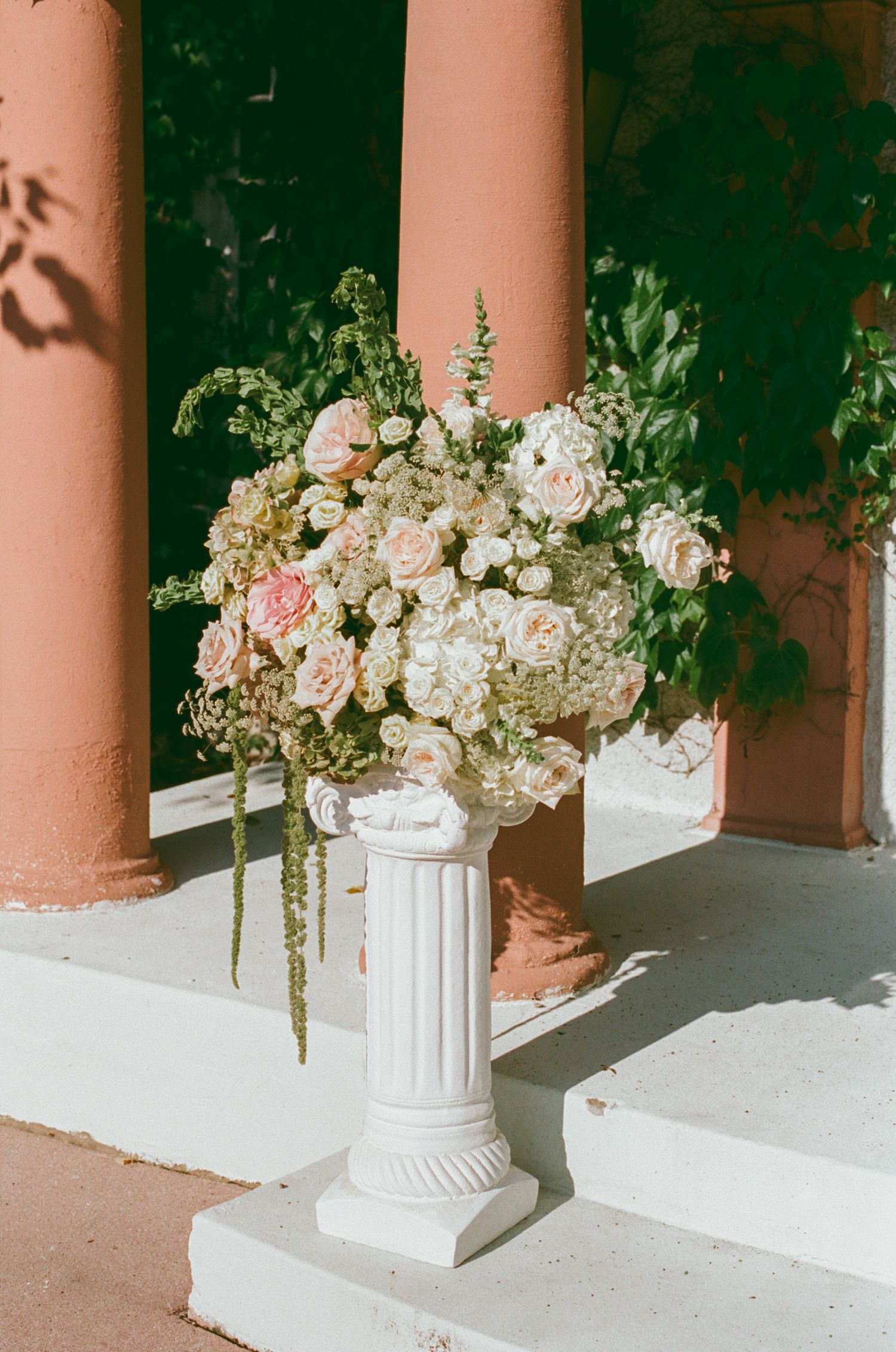 White pedestal vase with blush roses, hydrangeas, and greenery displayed between terracotta columns at an elegant wedding venue.