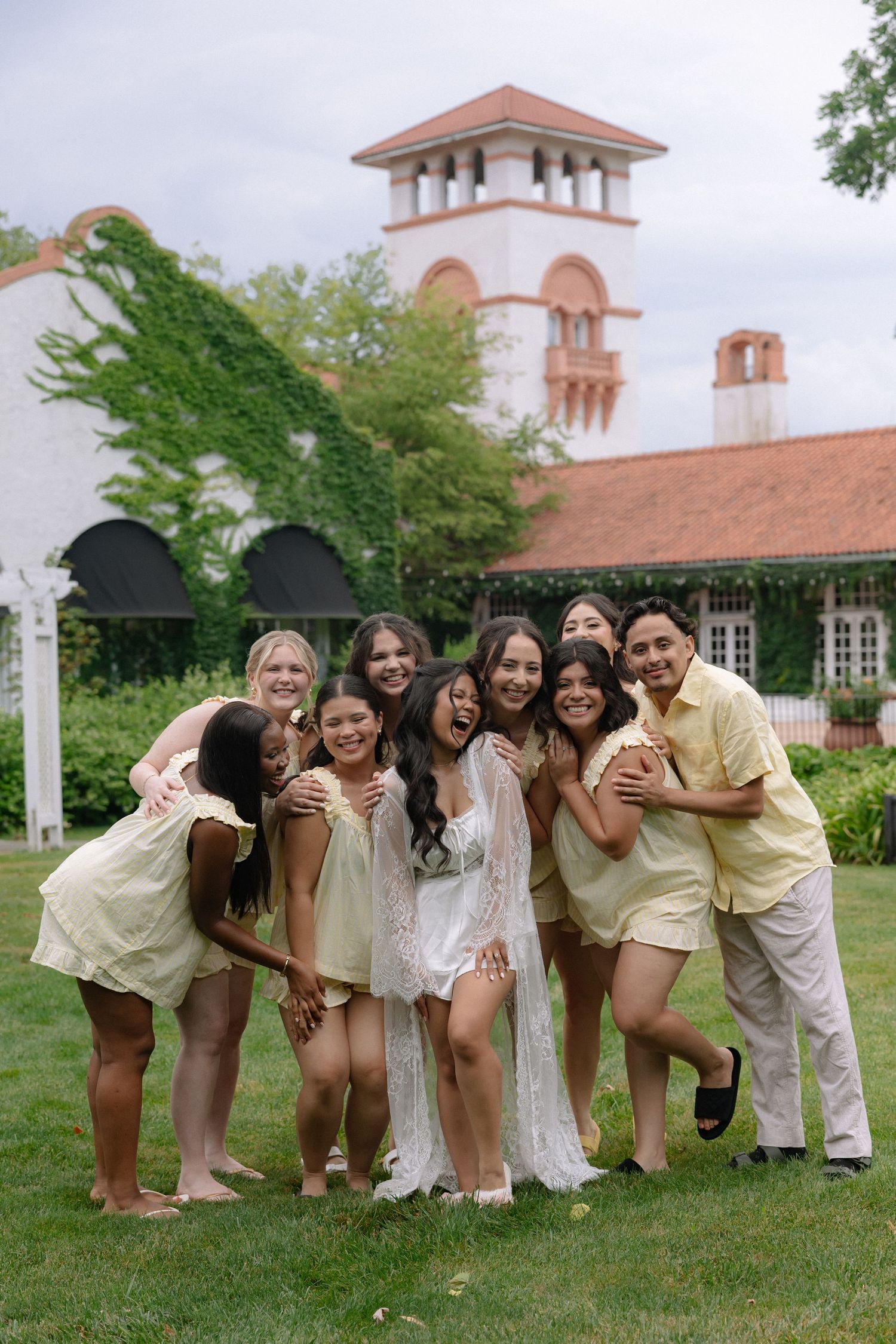 Bride in white lace robe laughing with bridesmaids in yellow dresses outdoors at elegant venue.