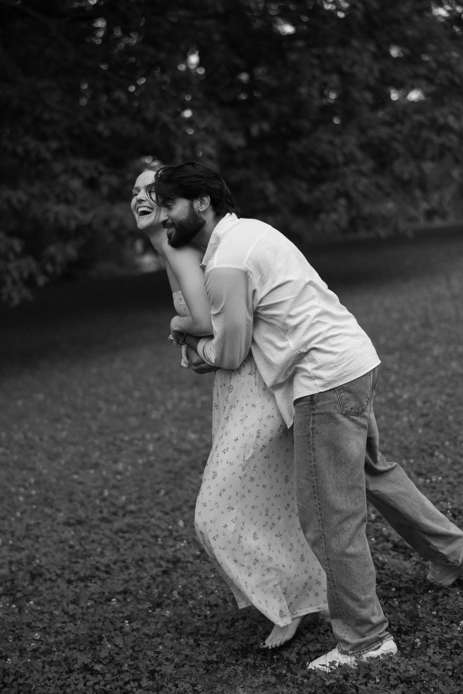 Black and white photo of a laughing couple embracing playfully outdoors in a lush green park setting.