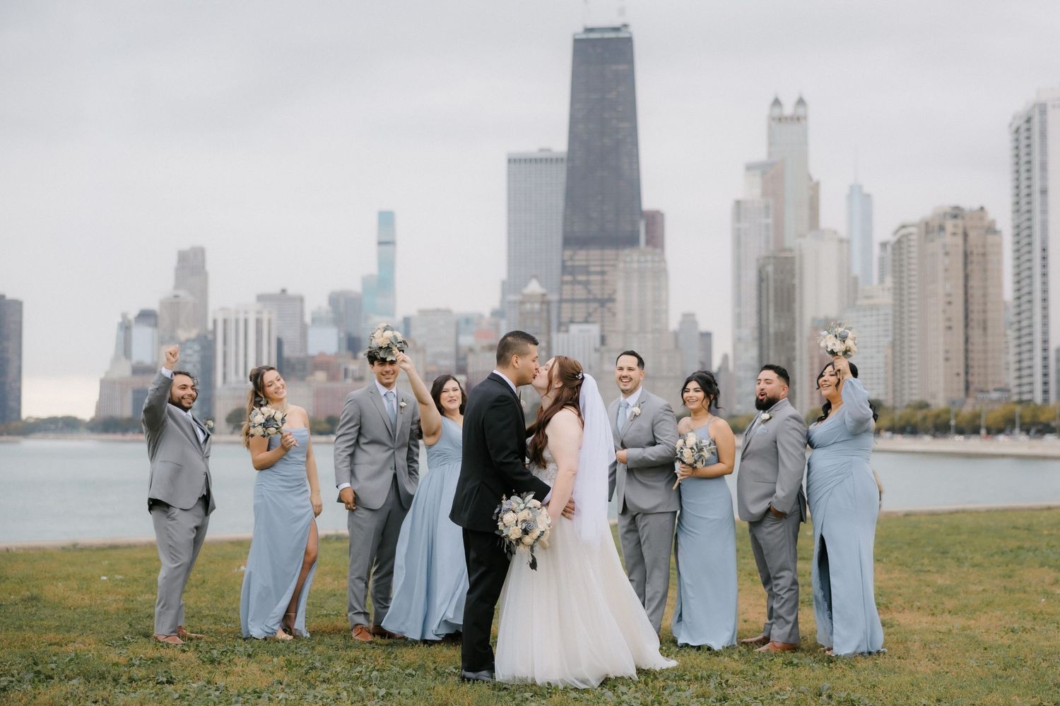 Wedding party poses with Chicago skyline backdrop near lakefront on cloudy day.