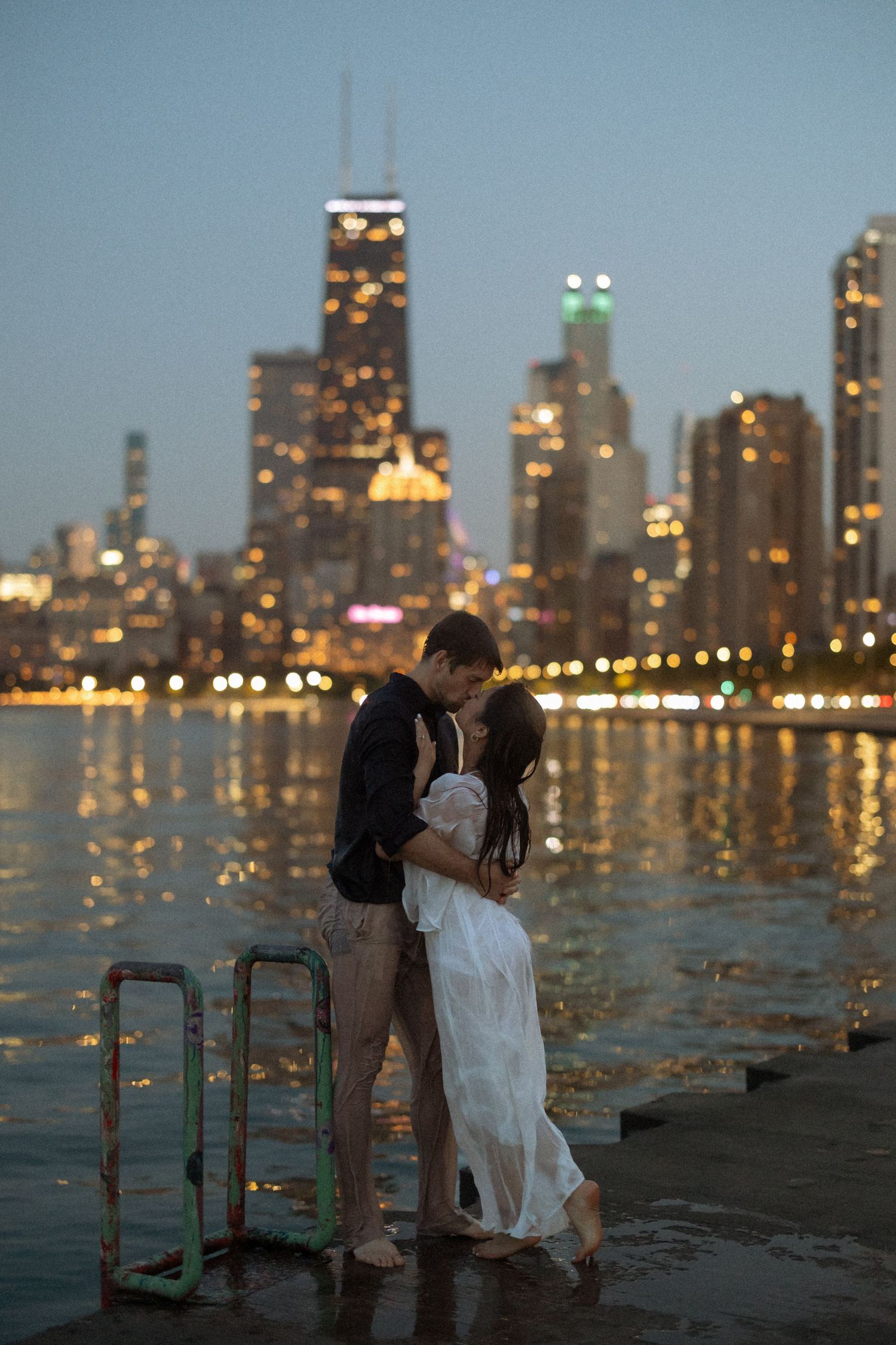 Couple shares romantic kiss on waterfront pier at night with glowing Chicago city skyline behind them.