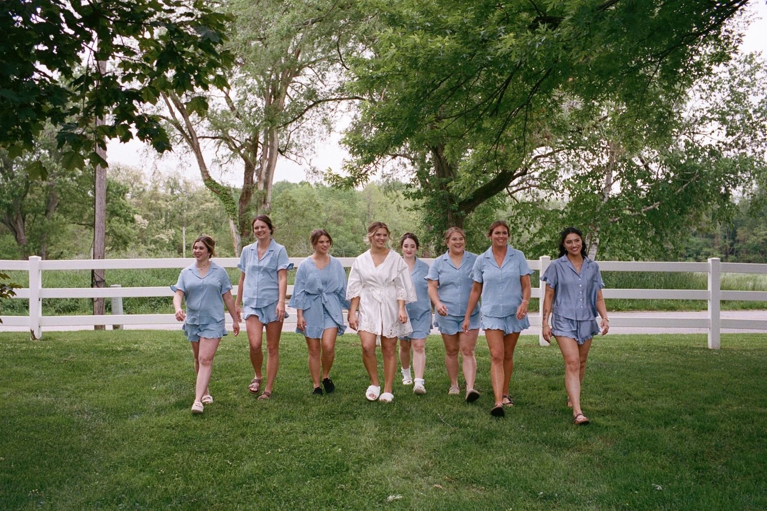 Bridal party in matching blue pajama sets walking together on green lawn with white fence and trees.