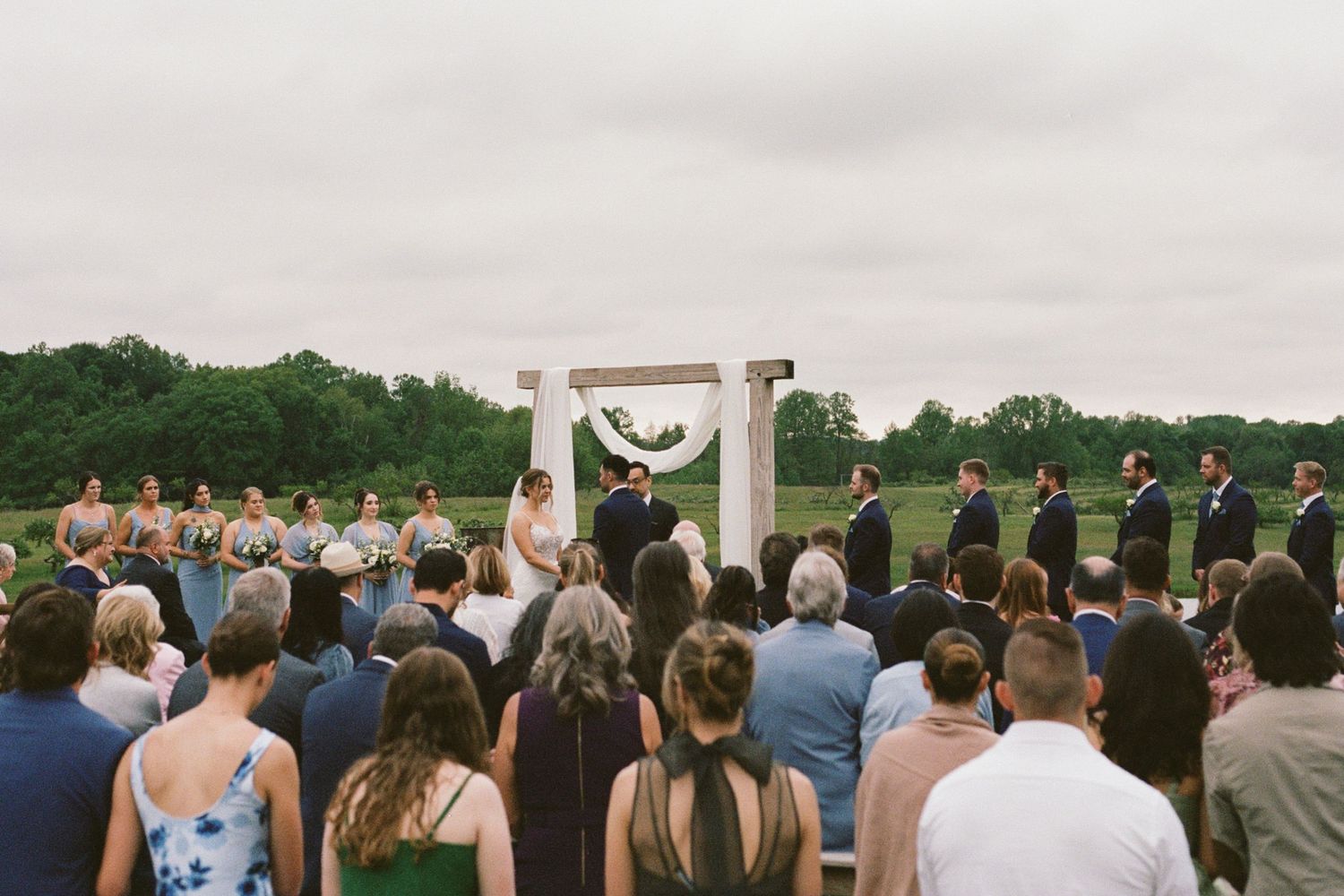 Outdoor wedding ceremony with couple at floral arch, bridesmaids in blue, groomsmen in black, guests standing on green lawn.