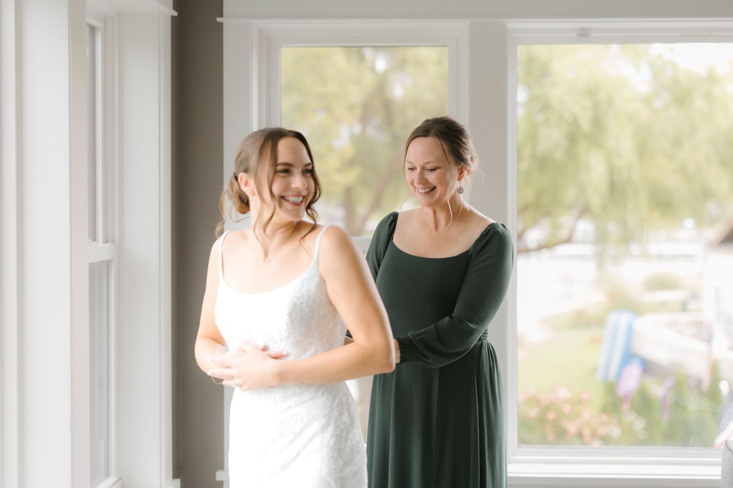 Bride in elegant white lace wedding dress smiles while woman in green dress fastens gown by window.