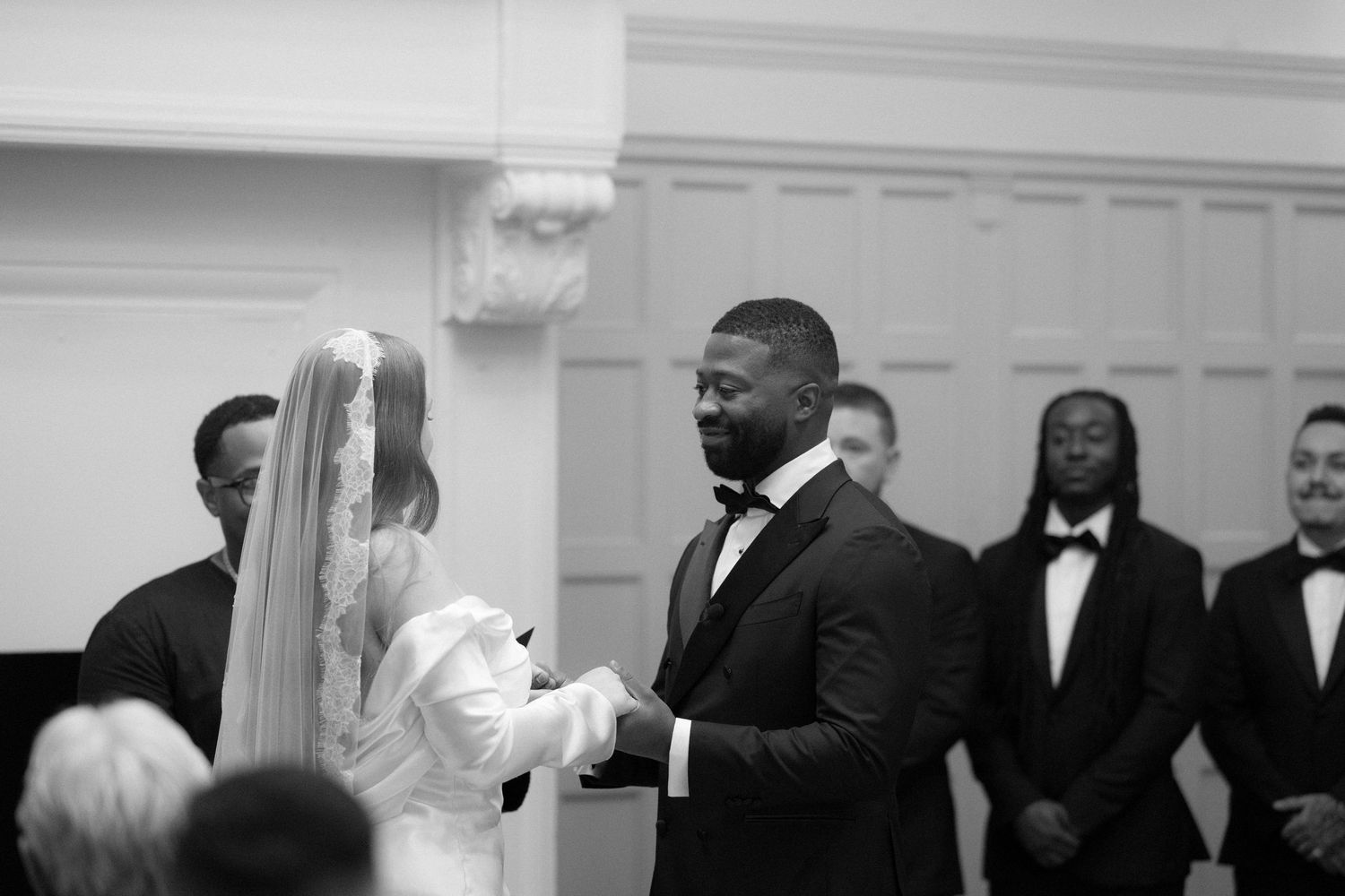 Black and white wedding ceremony photo of groom in bow tie holding bride's hands at the altar.