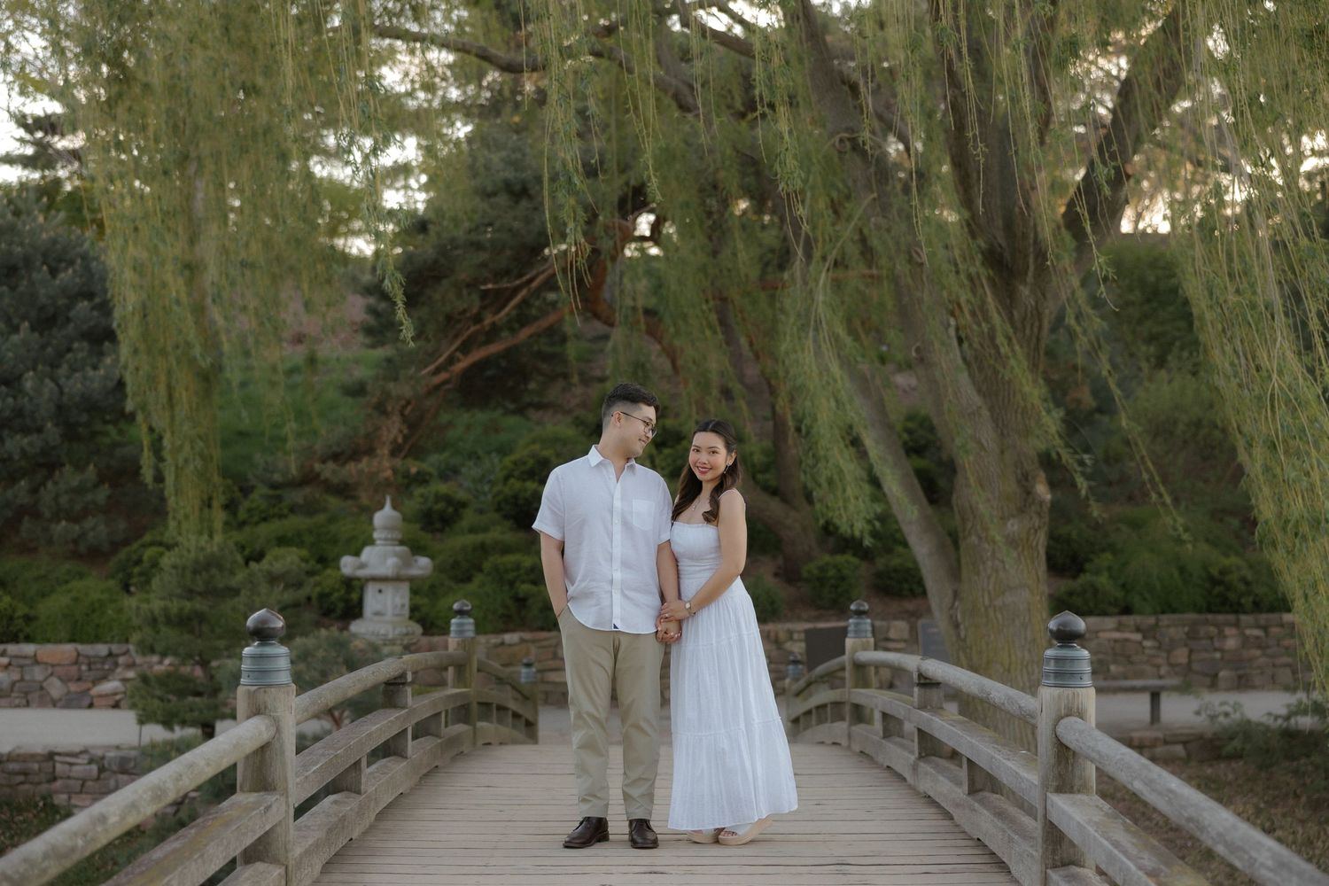 Couple standing on wooden bridge under weeping willow trees in Japanese garden during golden hour.
