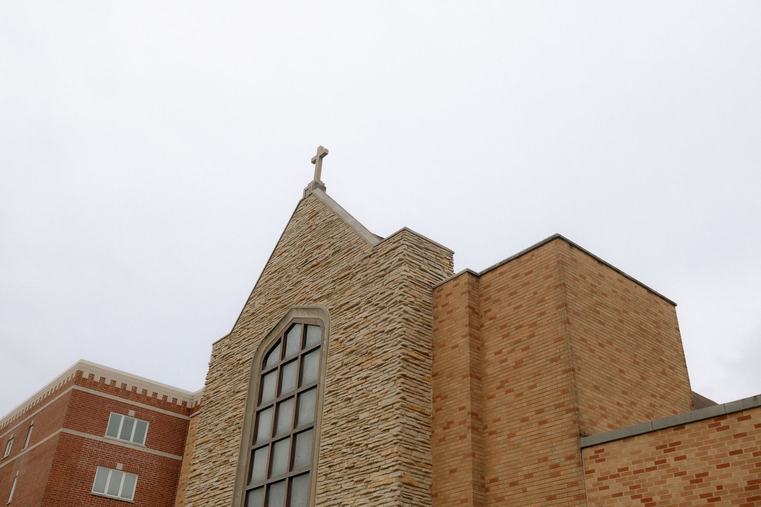 Stone and brick church exterior with arched window and cross atop pointed steeple against overcast sky.