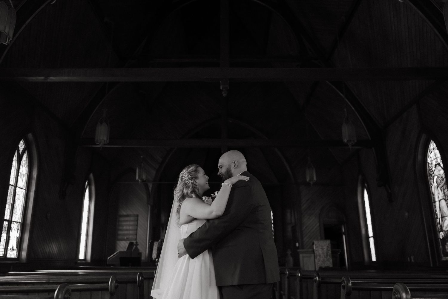 Black and white photo of a bride and groom sharing an intimate moment inside a historic church with arched windows.