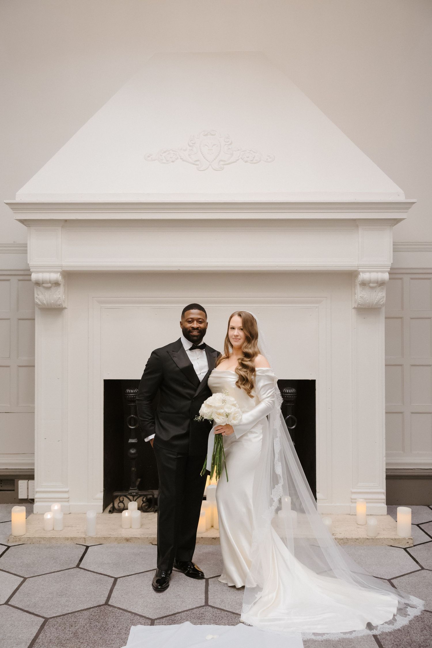 Interracial couple posing in wedding attire before elegant white fireplace with candles, bride holding white bouquet.