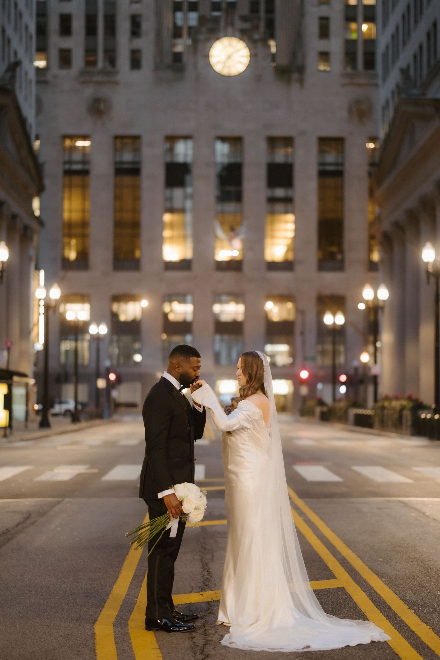 Bride and groom share a romantic moment on a city street at night, with a glowing clock tower backdrop.