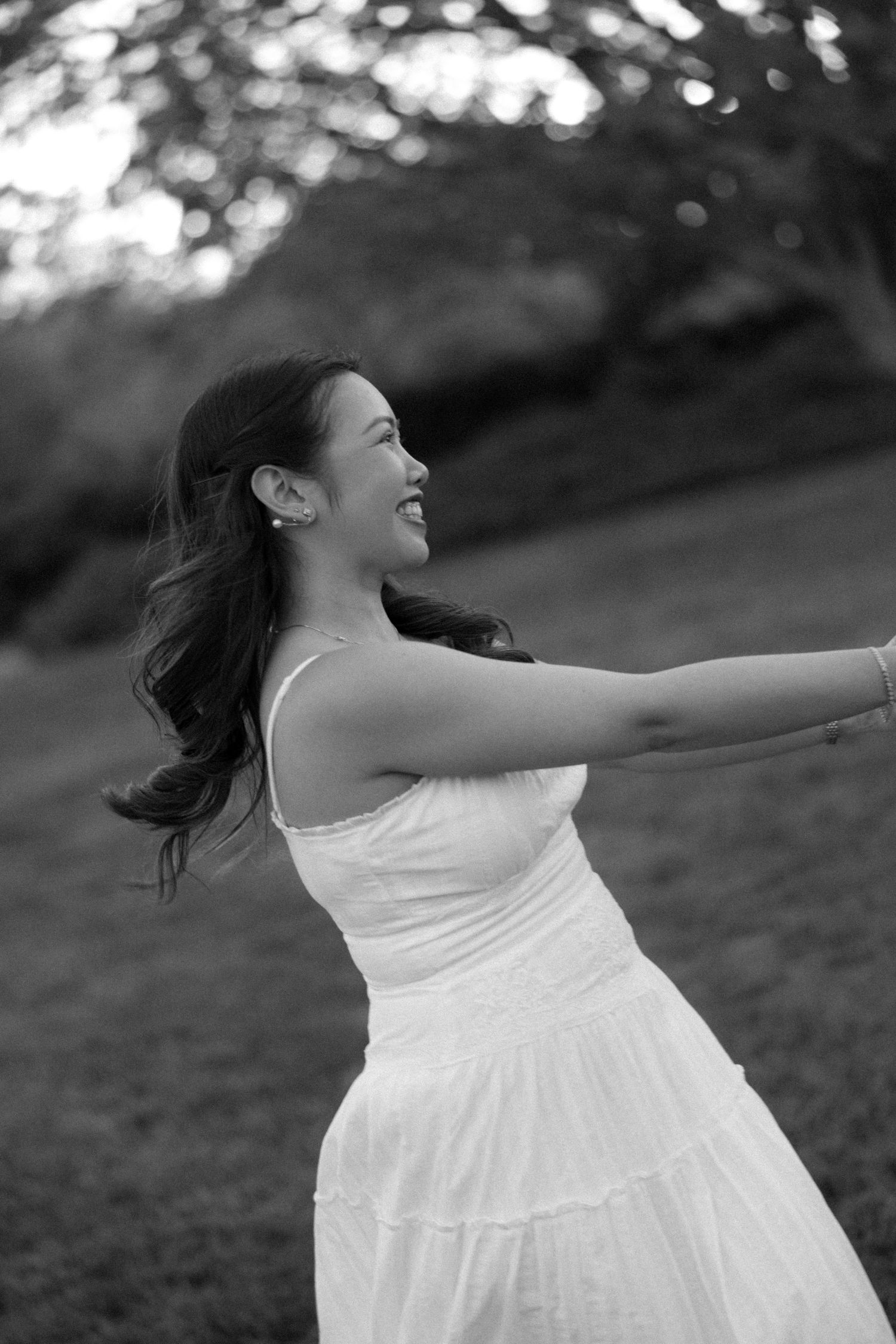 Black and white photo of a joyful young woman in a white dress spinning outdoors with arms outstretched.