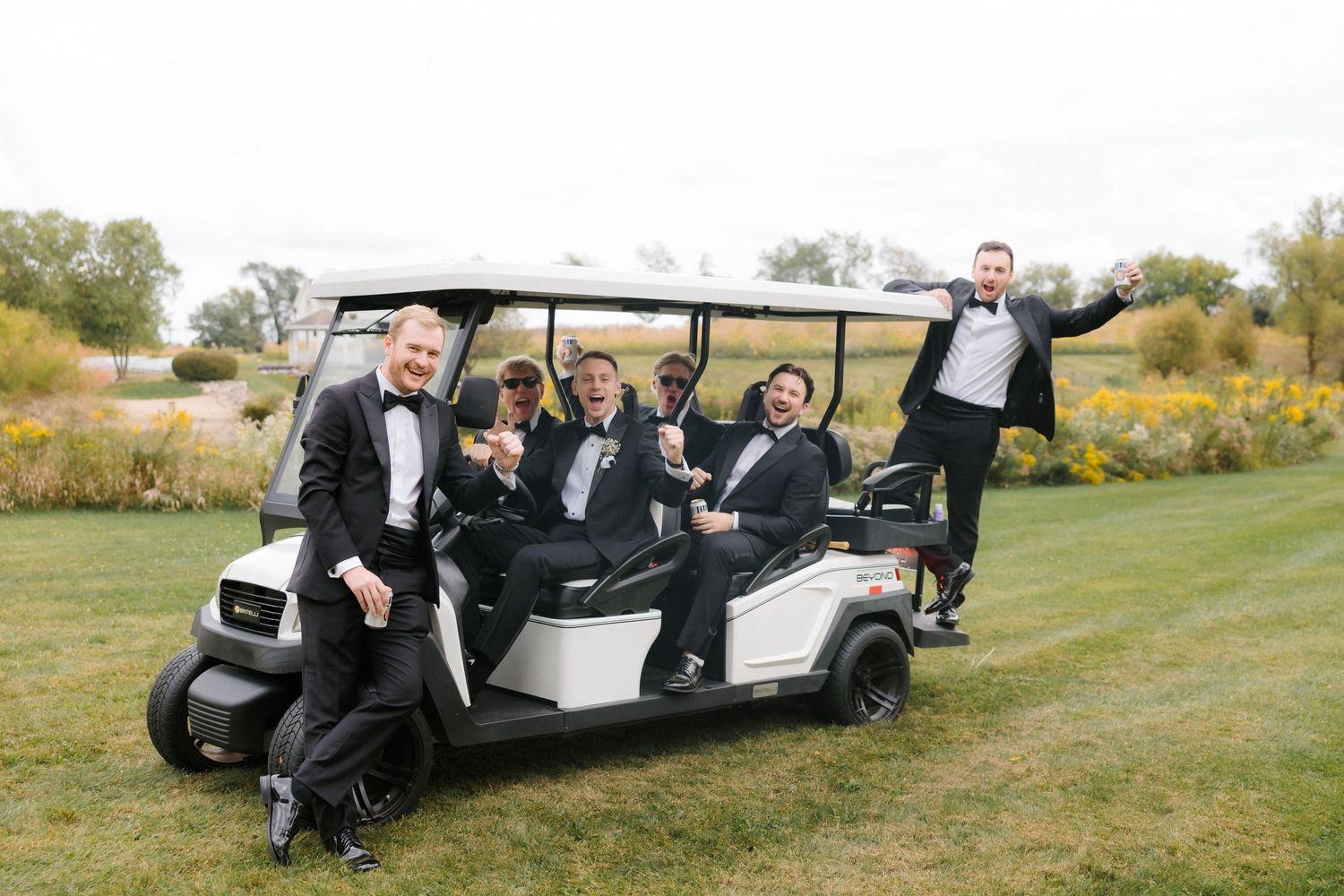 Groomsmen in tuxedos posing on a white golf cart on a green lawn at an outdoor wedding.