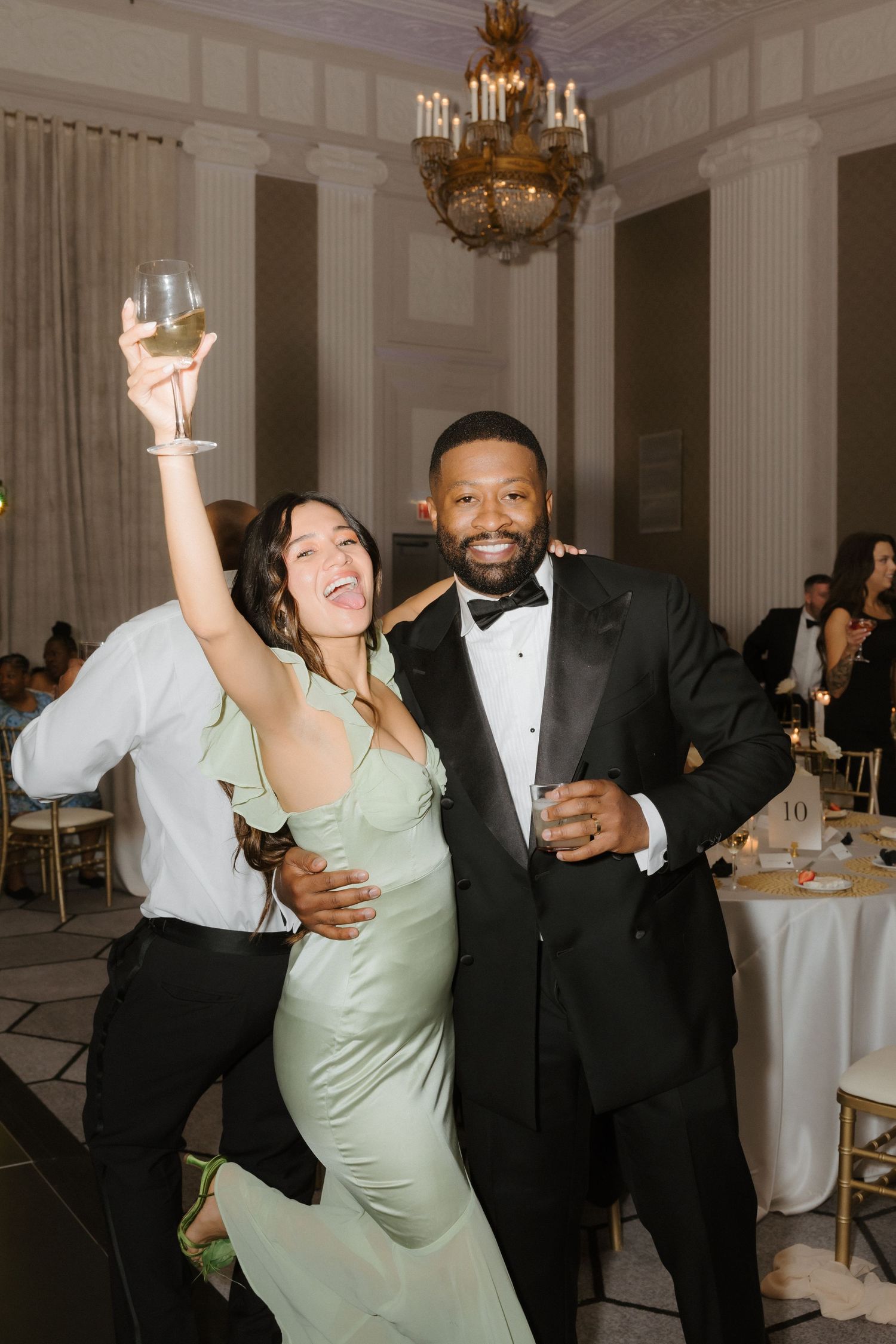 A woman in a mint green gown raises a wine glass while posing with a man in a black tuxedo at an elegant wedding reception.