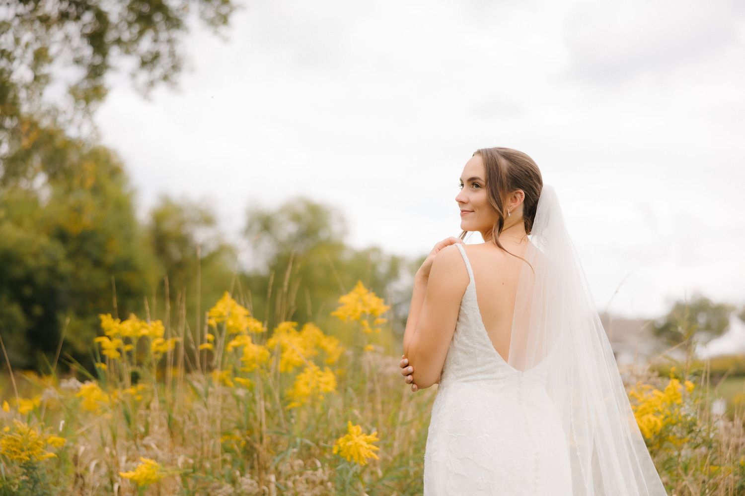 Bride in fitted lace wedding dress with flowing veil looking away, surrounded by golden wildflowers outdoors.