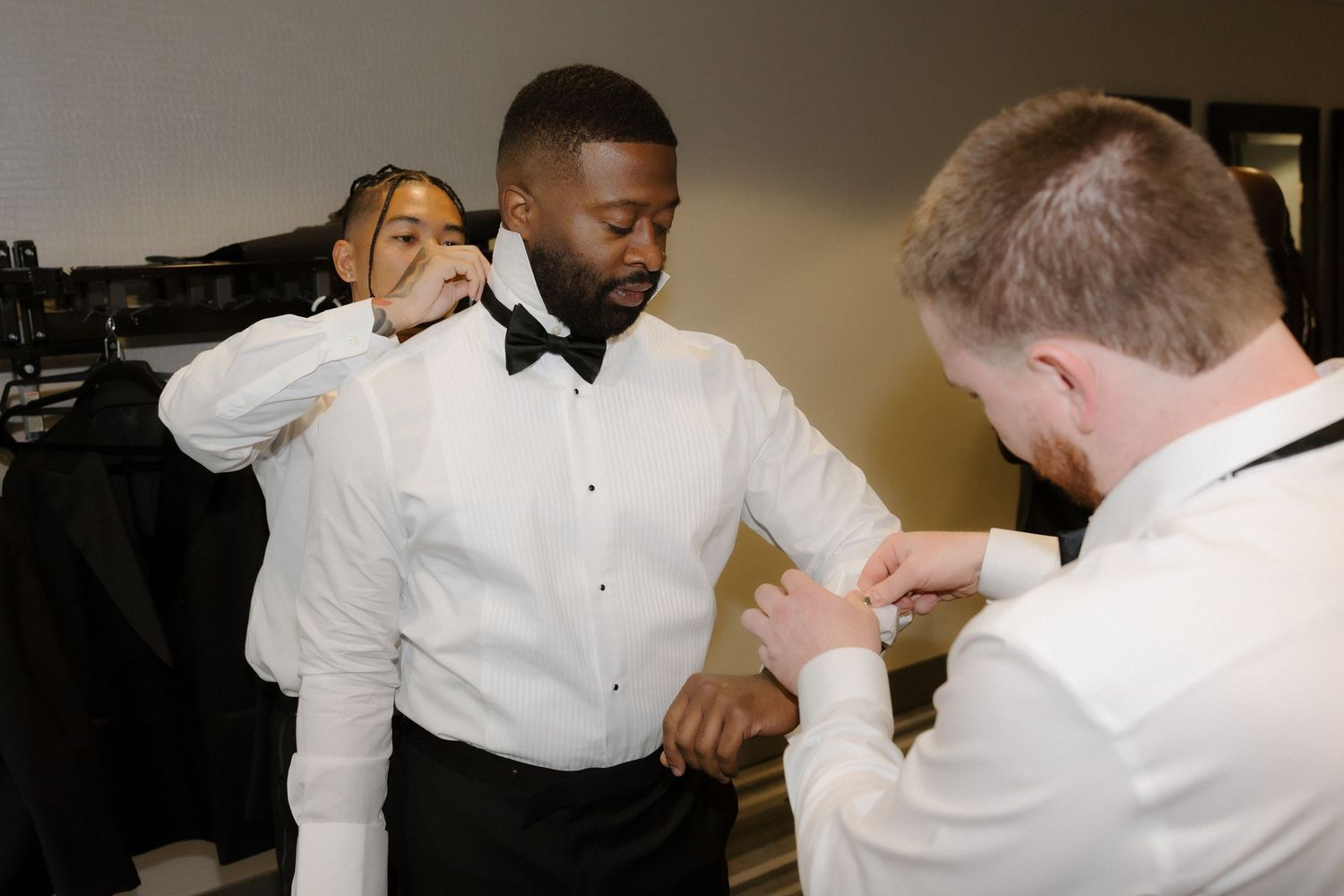 Three men in formal white dress shirts and bow ties helping each other get dressed for a wedding.