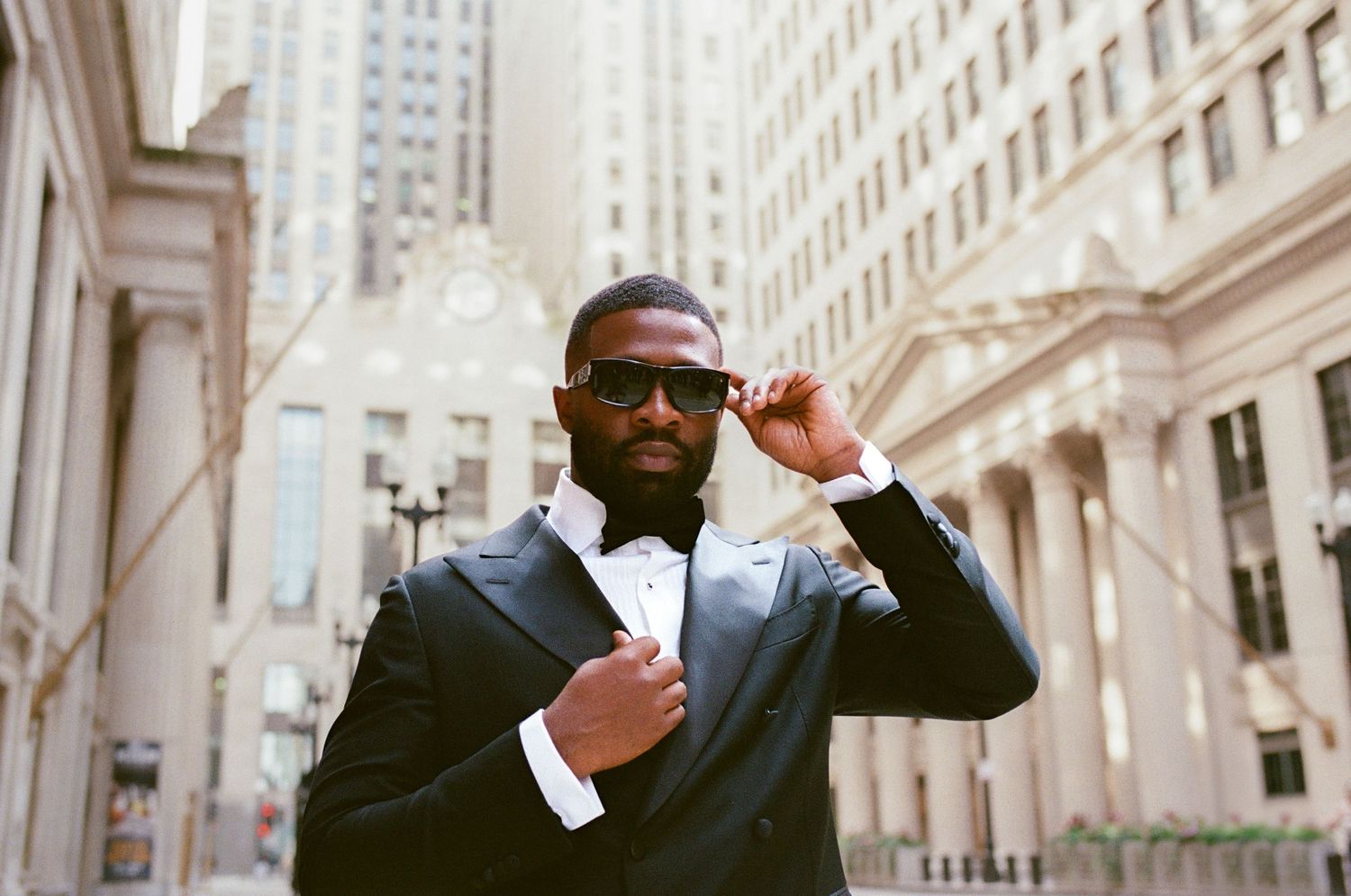 Stylish man in black tuxedo and bow tie adjusting sunglasses outdoors in urban city setting.