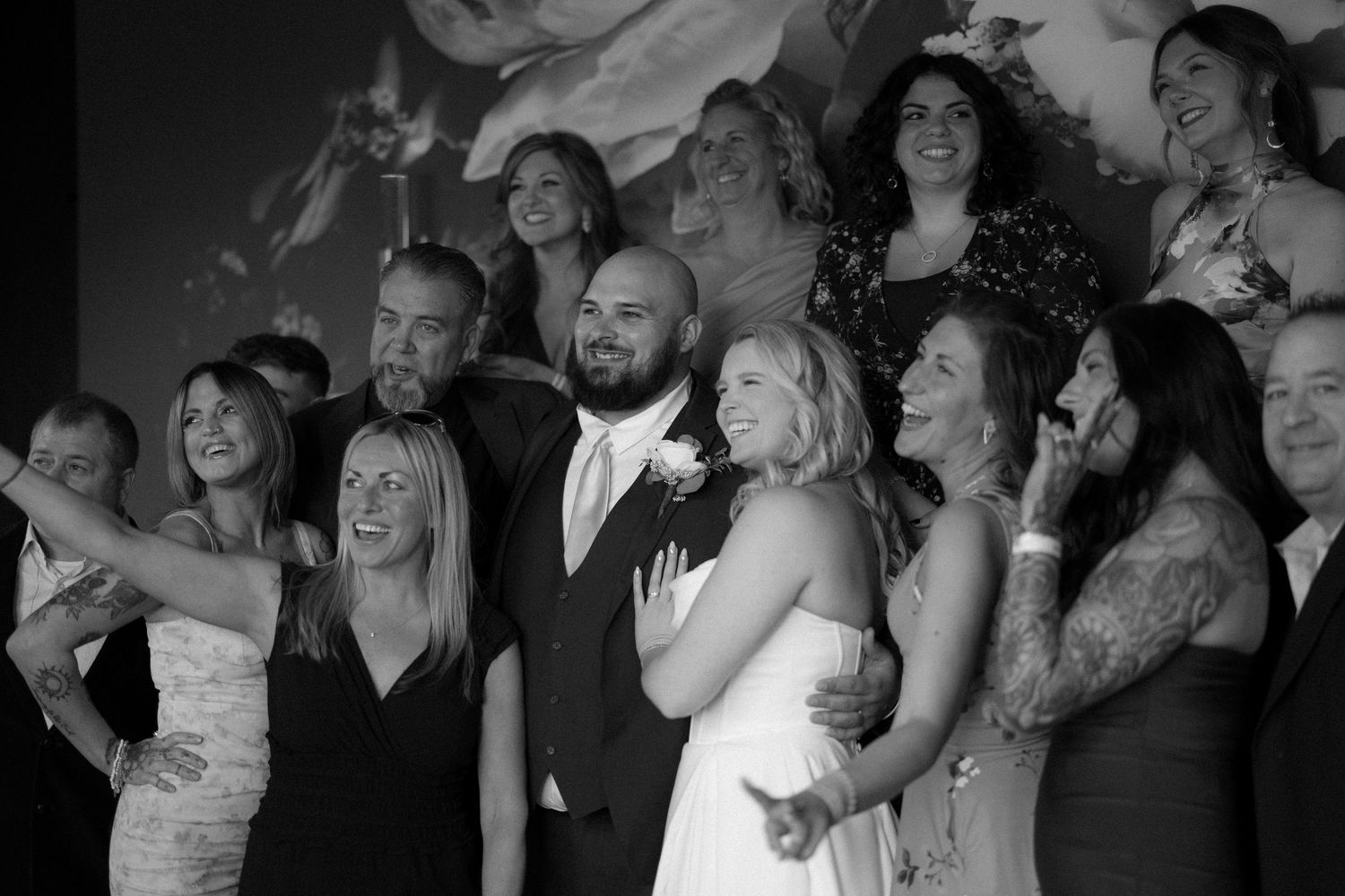 Black and white wedding photo of bride and groom posing with smiling guests at a formal reception.