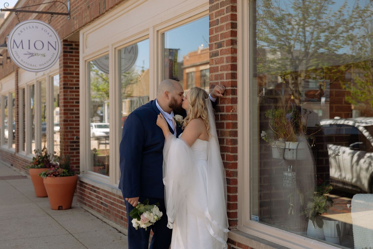 Bride and groom share a romantic kiss outside a brick storefront on their wedding day.