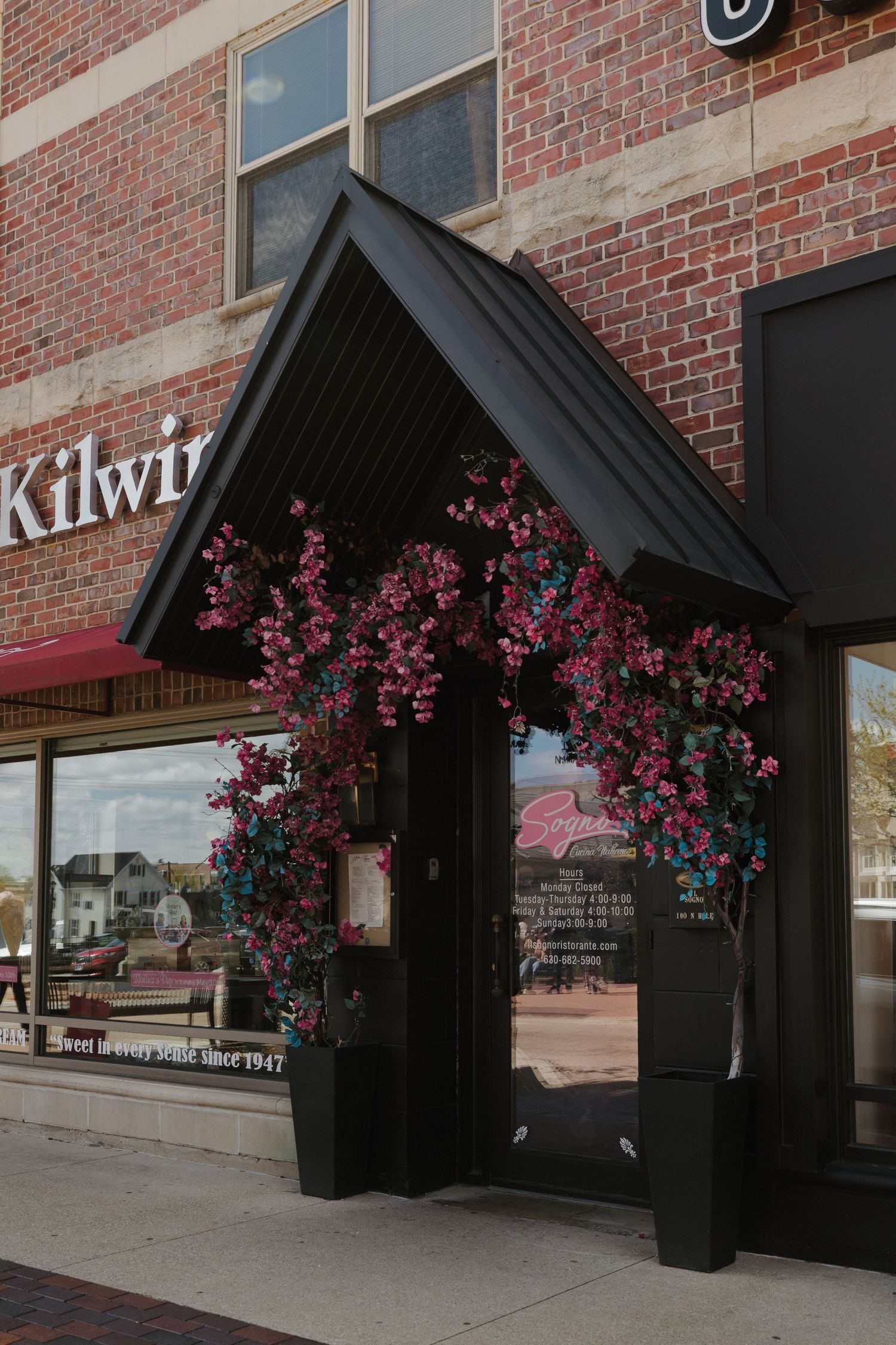 Black awning adorned with pink flowers at Kilwins chocolate shop entrance on a brick building streetfront.