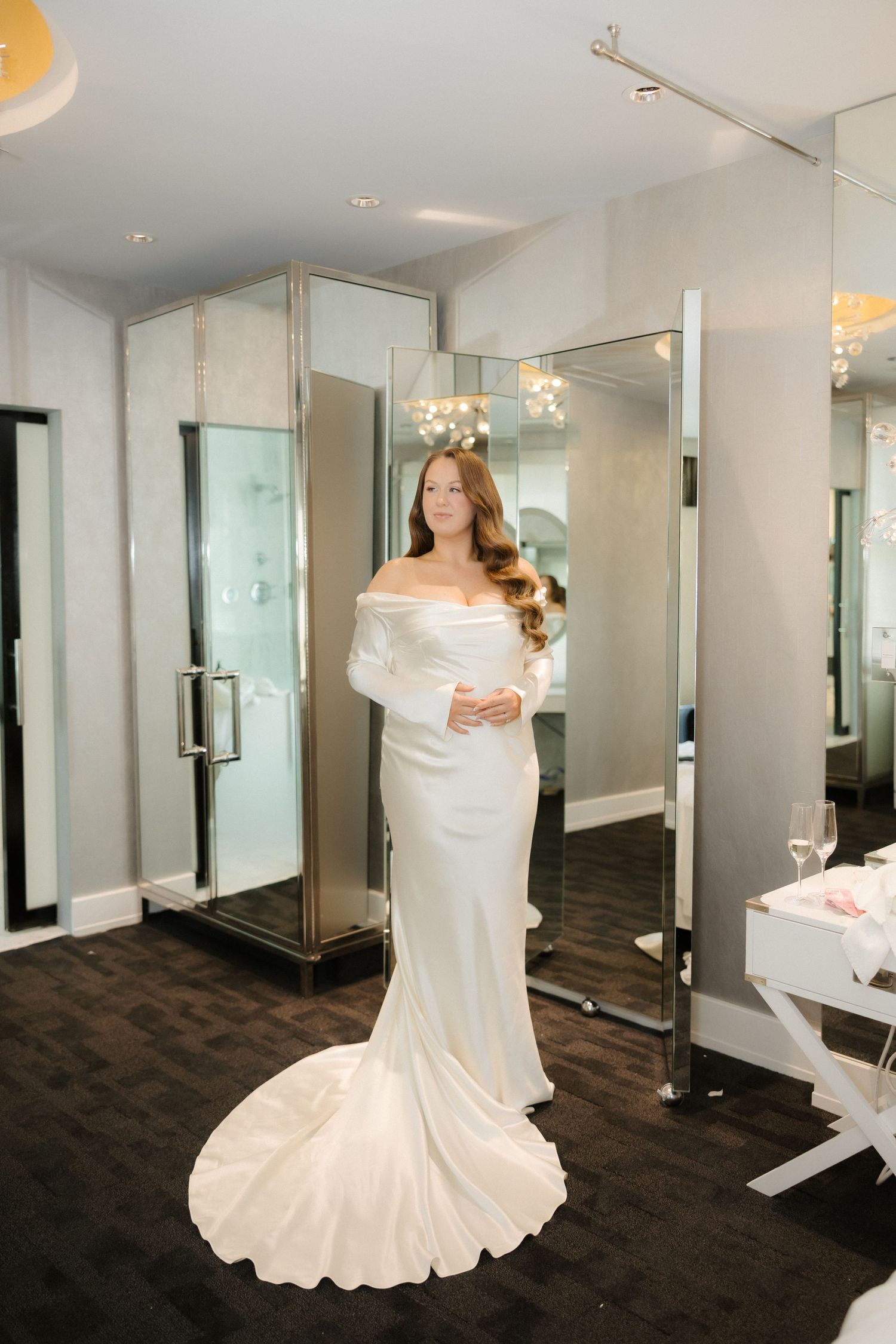 Elegant bride in white off-shoulder satin gown with long train posing in bridal boutique fitting room.