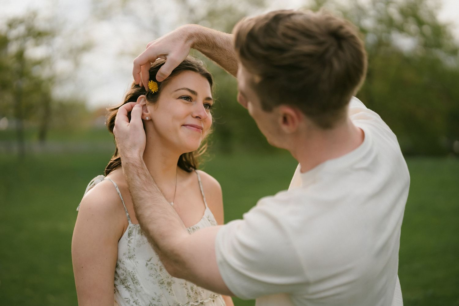 A man gently places a yellow flower in a smiling woman's hair in a green park.