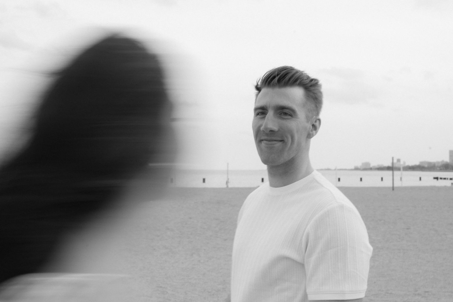 Black and white portrait of smiling man in white t-shirt near a beach.