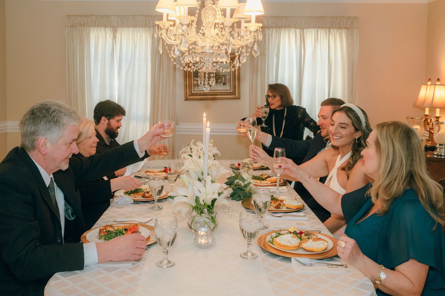 Wedding guests raising champagne glasses in a toast at an intimate dinner reception under a chandelier.
