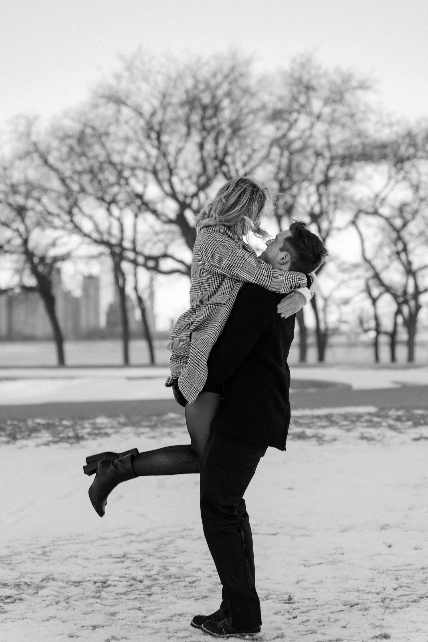 Black and white photo of man lifting woman in snowy park with bare trees and city skyline behind them.