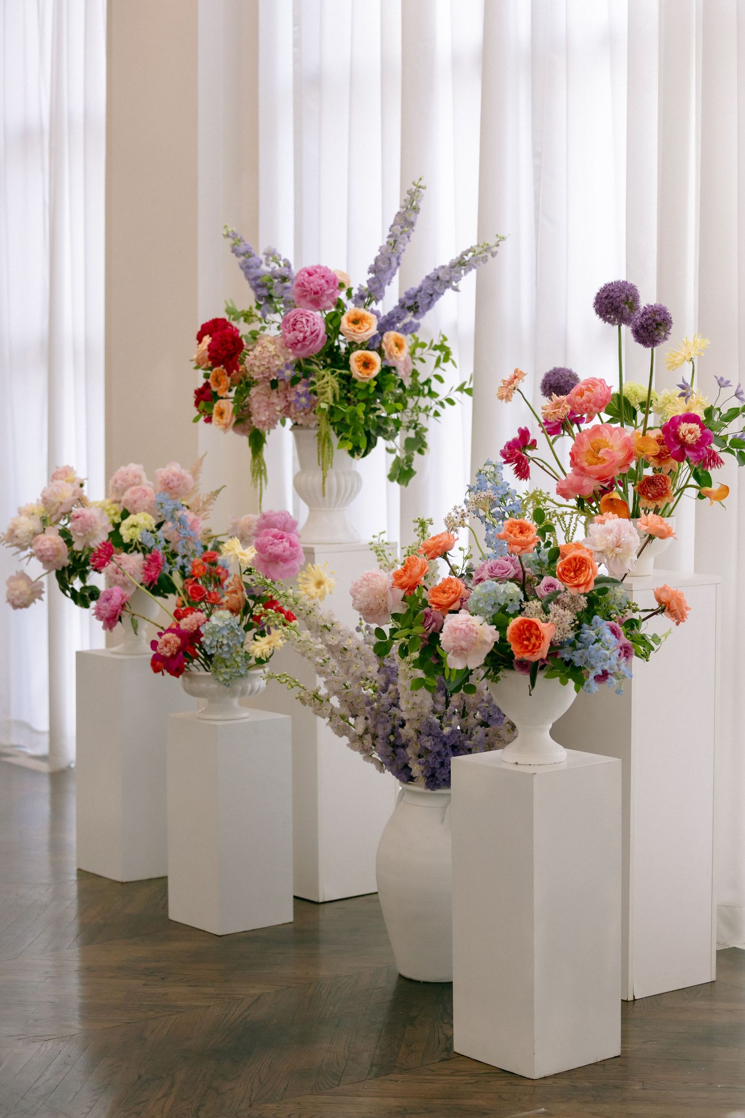 Colorful floral arrangements in white vases on pedestals featuring roses, dahlias, and delphiniums in a bright white room.