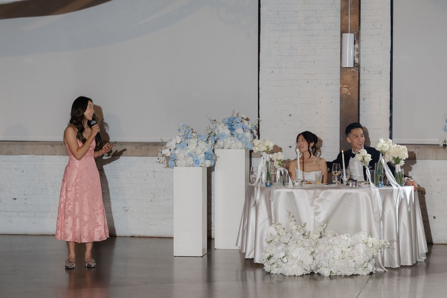 Woman in pink dress giving a speech at a wedding reception while the bride and groom sit at the head table.